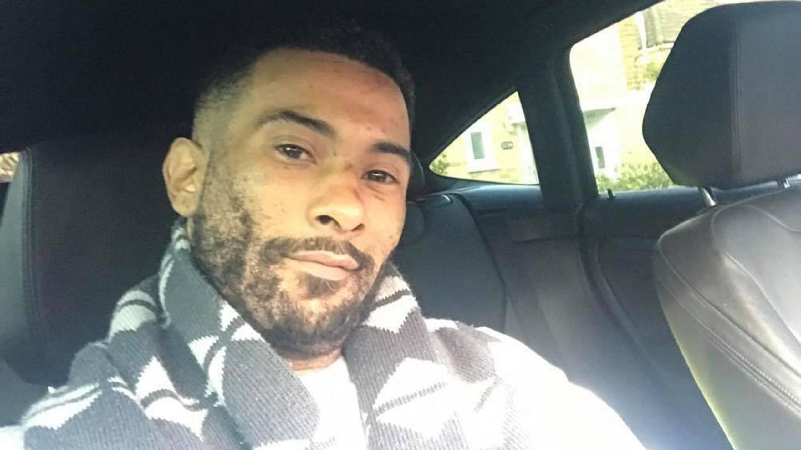 Leroy Mitchell is sitting in a car, smiling into the camera in a selfie-style photo. He has short black hair and a short black beard and is wearing a black and white scarf.