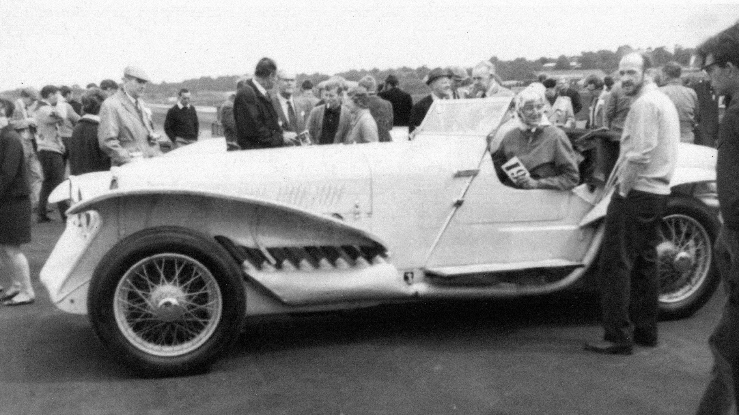 It is a black and white photo from 1922. It is of a Leyland Eight car. It is at a race track and there are people surrounding the car chatting and talking. The car is of a side view and is long in shape with a woman sat in the drivers seat