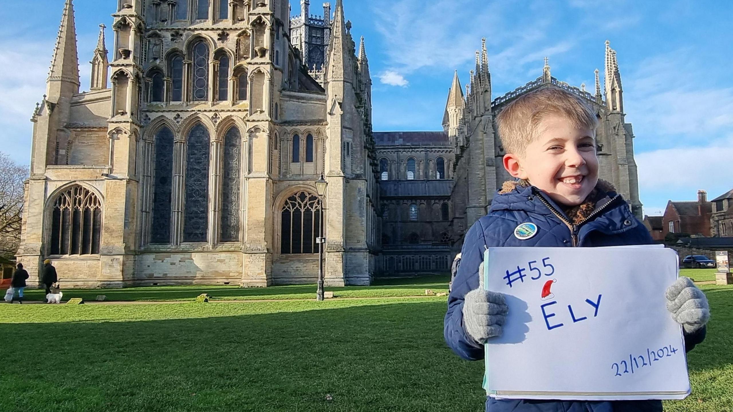 Austin is in a blue coat and grey gloves. He is looking at the camera and smiling and is holding a white sign which says #55 Ely, 22/12/2024. Behind him is Ely cathedral