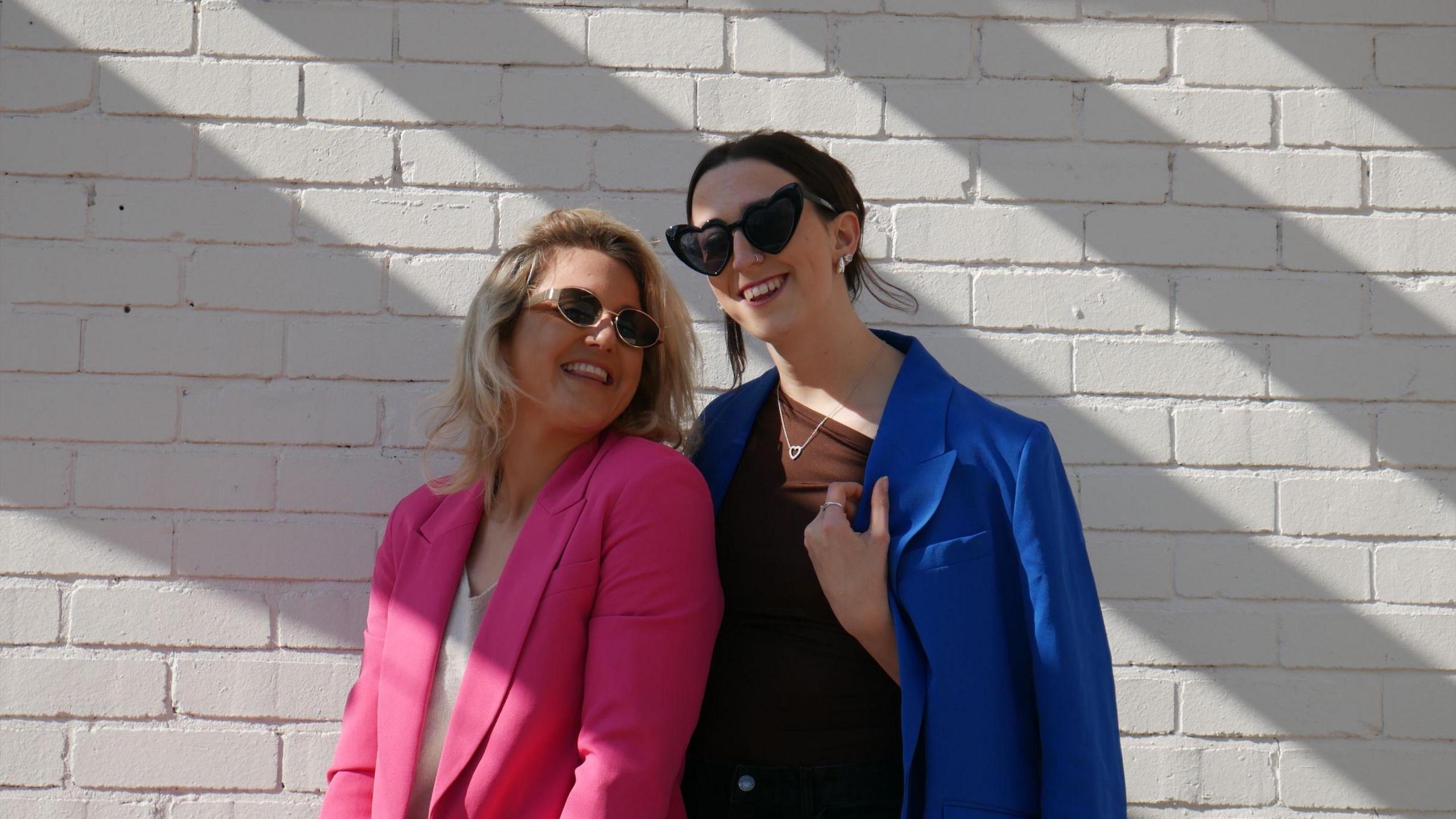 Two young women, one wearing a pink blazer and the other a blue blazer, stand together in front of a whitewashed brick wall wearing sunglasses.