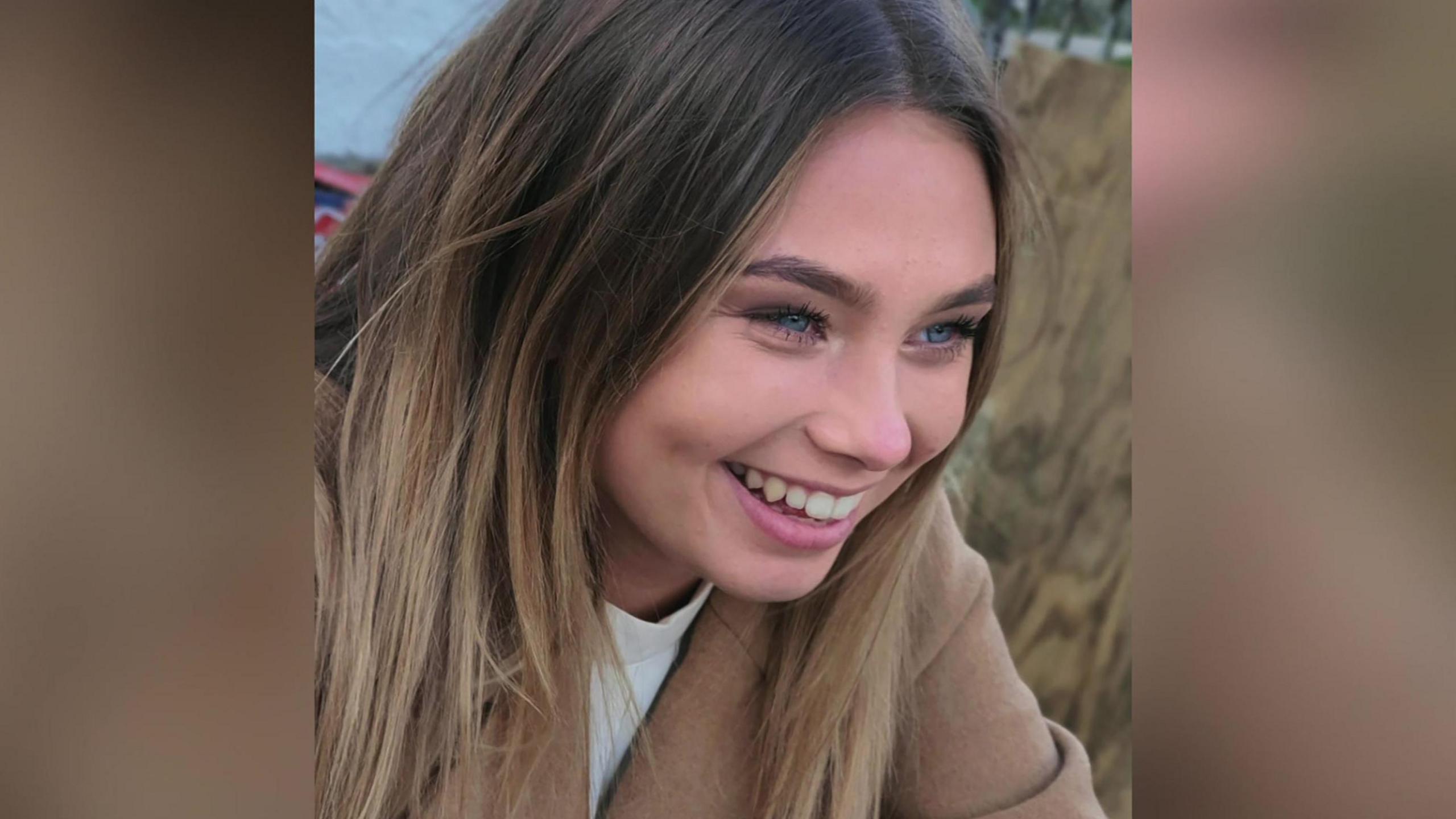 A teenage girl with blue eyes, and blonde hair wearing a brown jacket is pictured smiling. 