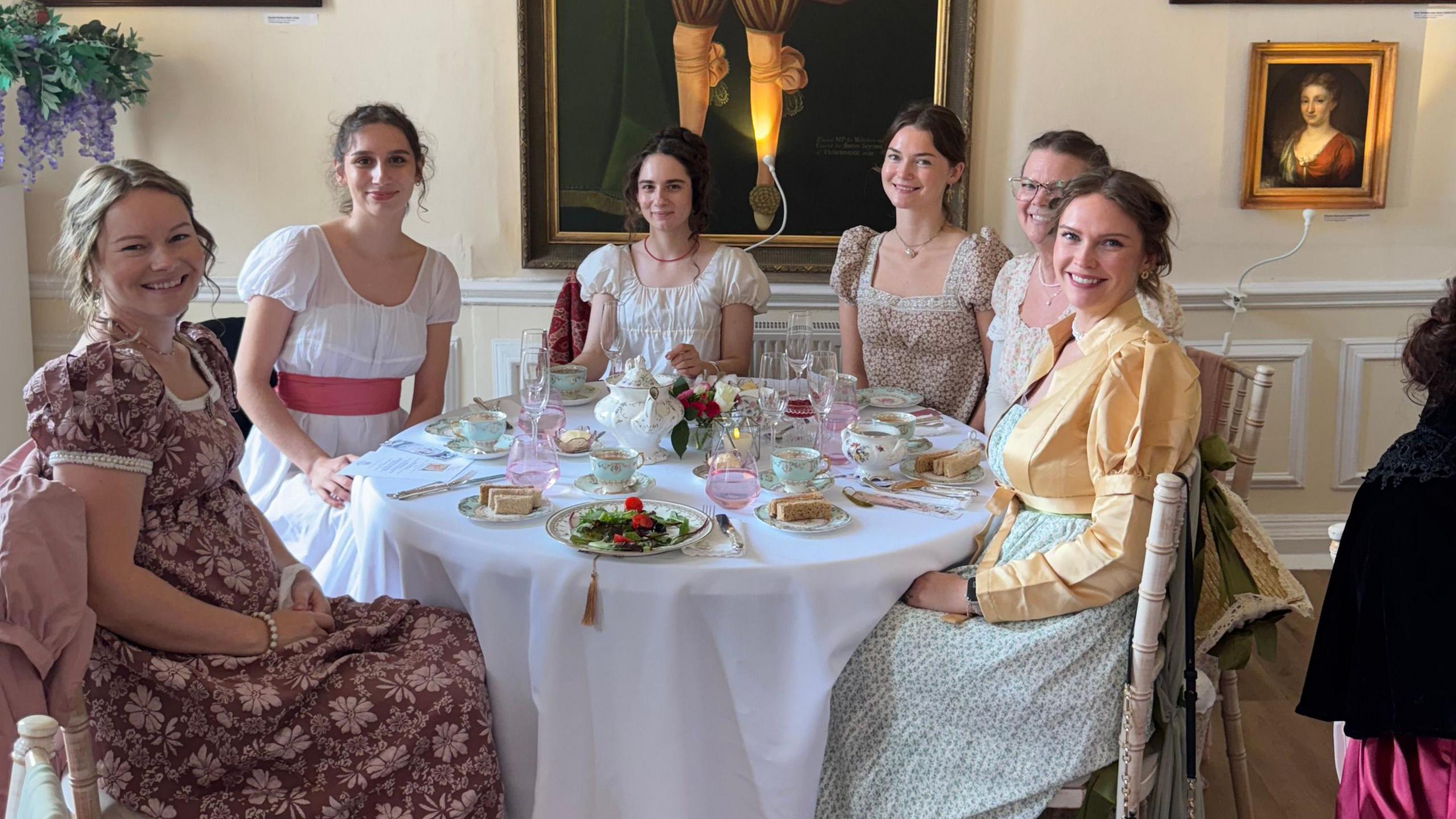 A group of women in Regency inspired dresses gathered around a round table with a white tablecloth, old china and small sandwiches and cups of tea.
