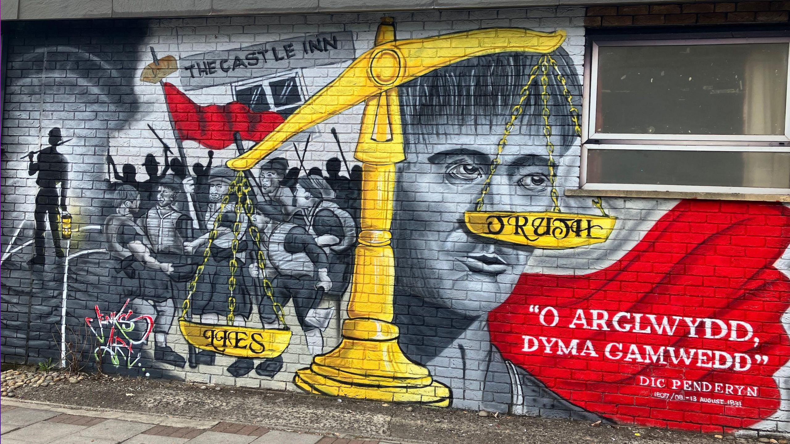 Brick wall with black and white graffiti depicting the face of Dic Penderyn, the scene of the Merthyr Rising with a number of people holding sticks in the air and a red flag. There's also a large golden scales in the centre of the mural with the words Lies and Truth written each side. On the lower right side of the mural is a block of red with the words "O Arglwydd dyma gamwedd" ("Oh Lord this is iniquity") which are believed to be the final words uttered by Dic Penderyn before he was executed.