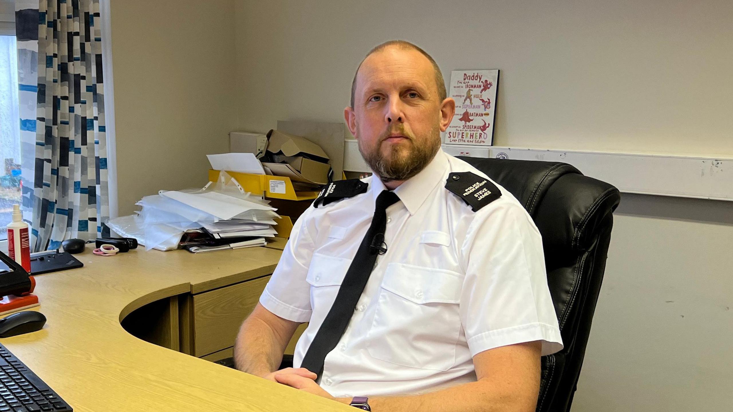 Sergeant Steve James pictured behind his desk in his office. He is wearing uniform and has a light brown beard. 