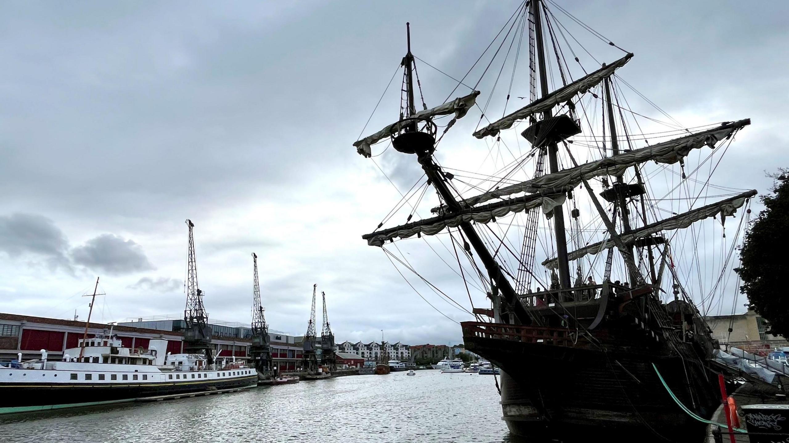 A pirate-style dark coloured ship called the Galeón AndalucÃa is seen moored in Bristol's harbour, with the cranes of the MShed museum visible in the background.