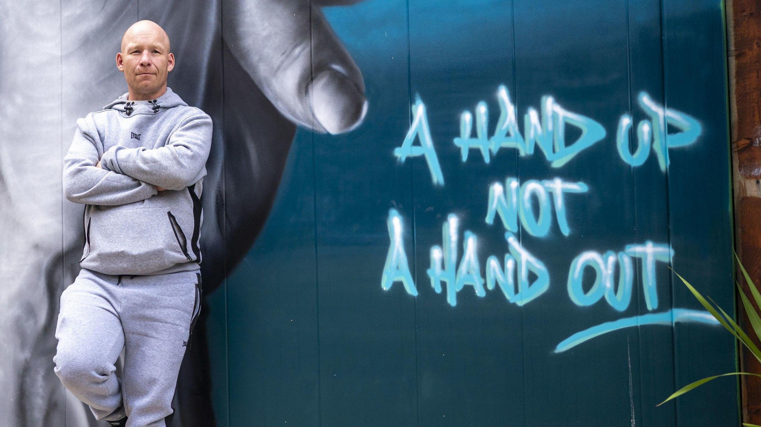 A man in a grey tracksuit top and bottoms leans back against a wall which has street art on it. Behind in is a large pair of hands - painted in grey - intertwined and then there is a dark green wall which has written on it "A hand up not a hand out"