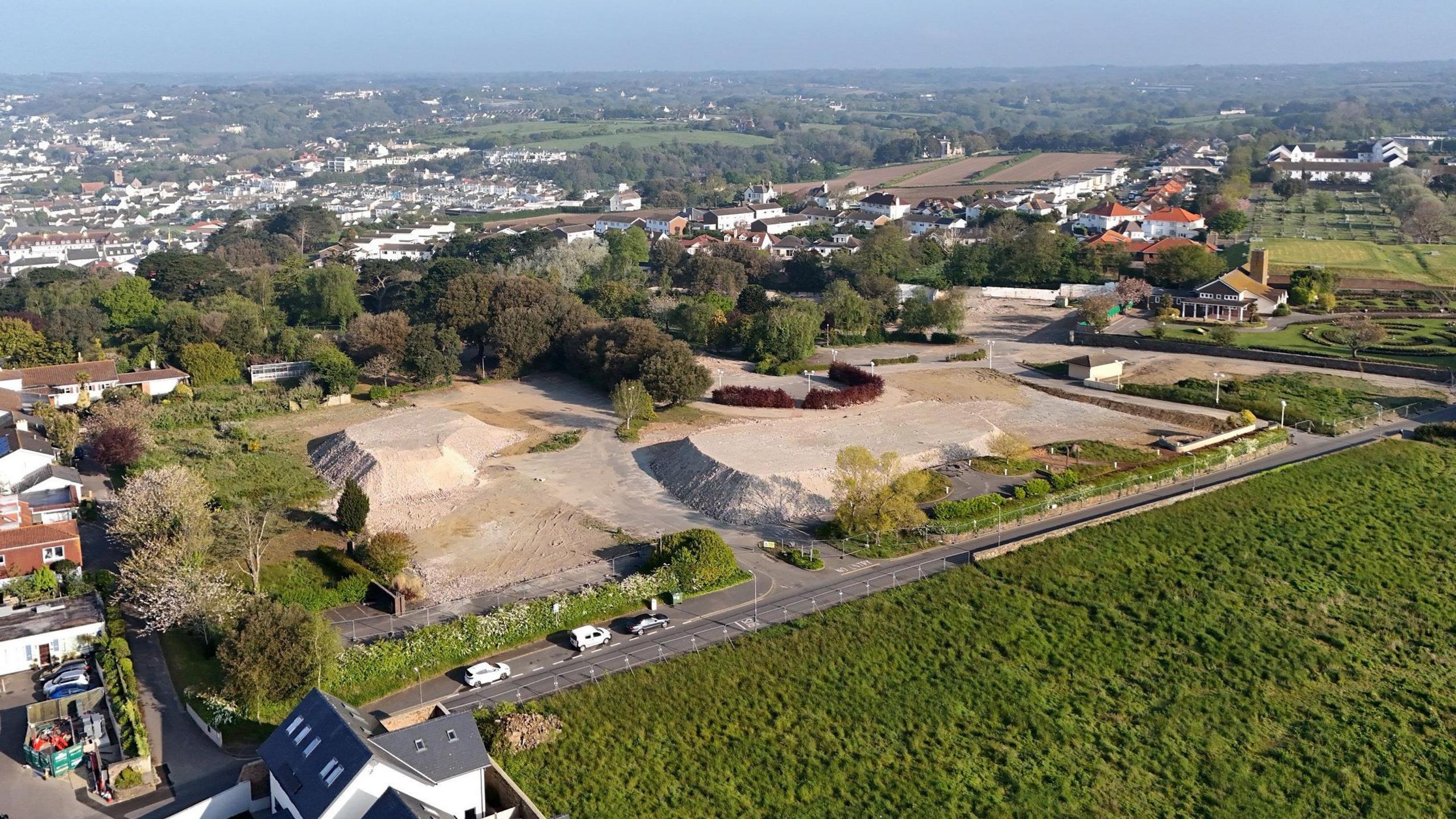 Drone shot overlooking the Overdale Acute Hospital development site in St Helier. Large mounds of building material are at the site. The area is surrounded by trees, fields and buildings.