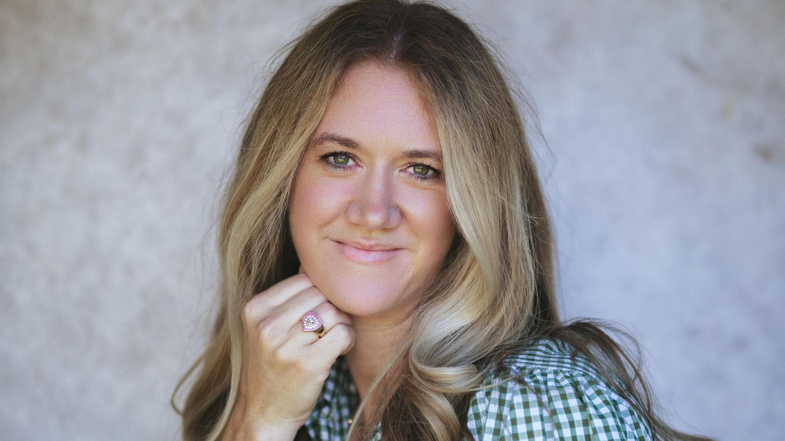 A woman with long blonde/brown hair smiles to the camera. Her right hand is resting under the right Jaw. She has a pink heart ring on her ring finger. She wears a green and white chequered shirt. 