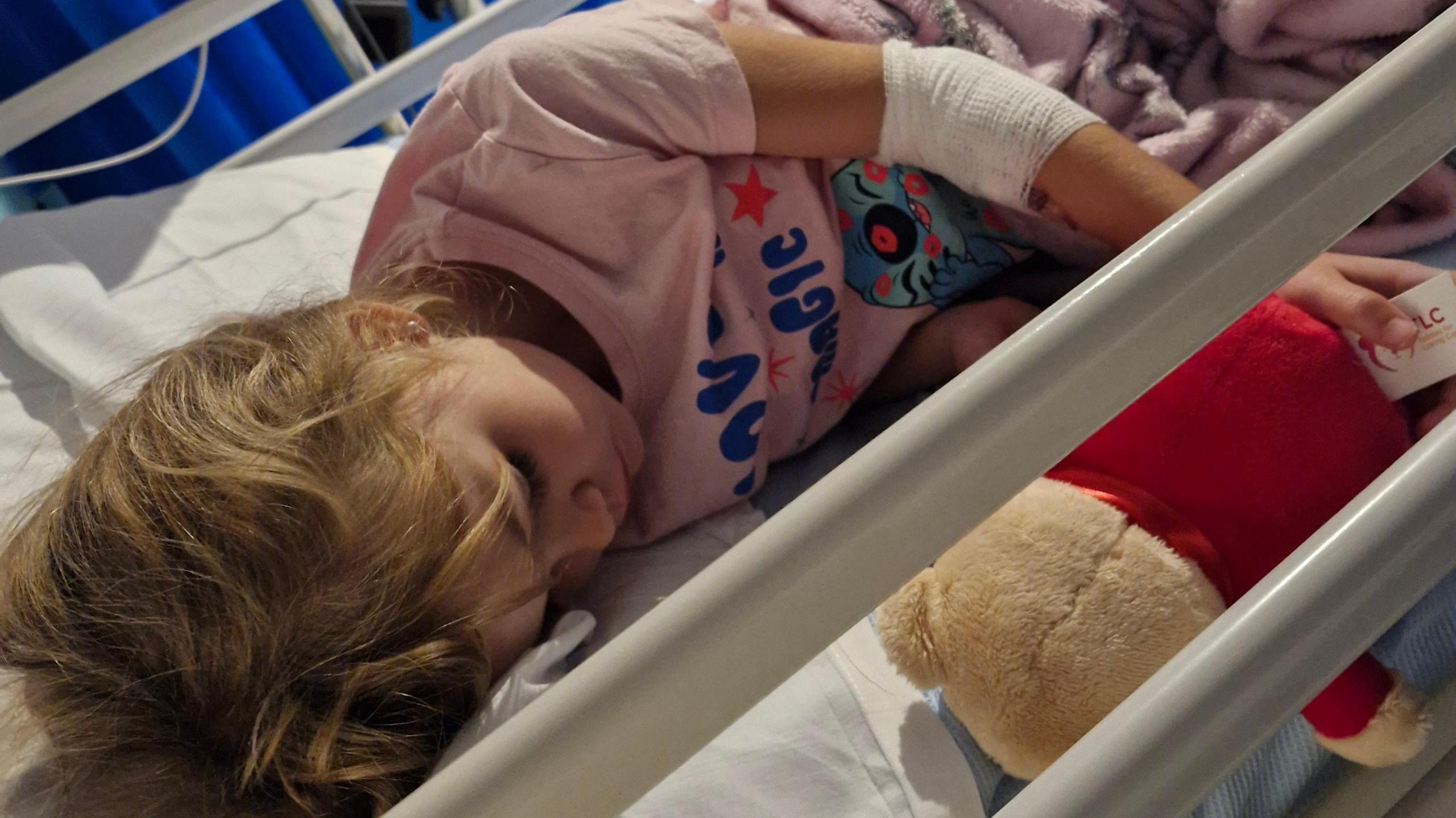 A young girl wearing Lilo and Stich pyjmas is laying in a hospital bed with a Winnie-the-Pooh teddy bear next to her.