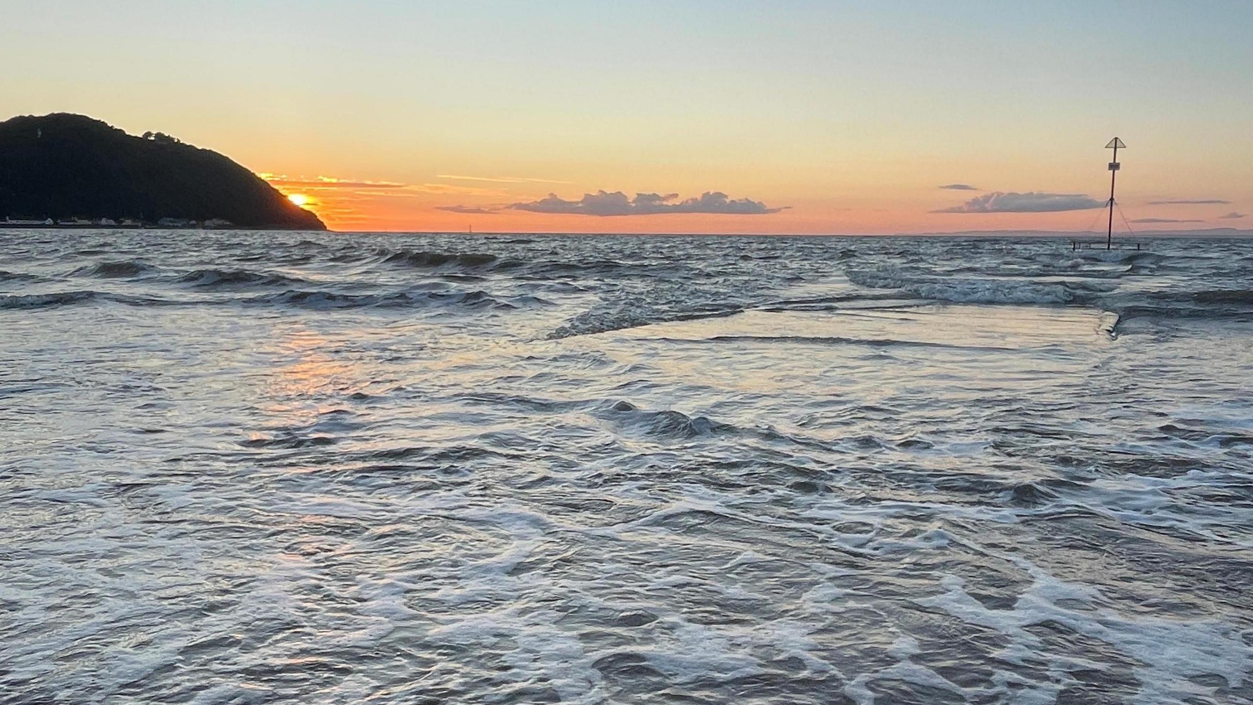 The sun is setting over the sea, which is white and foaming as it comes in over the sand. There is a hill in shadow to the left.