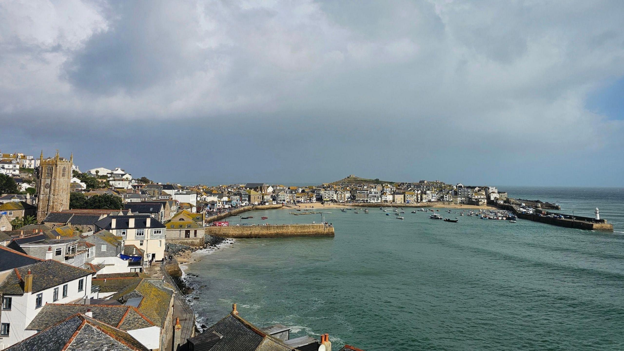 A picture looking down on the town of St Ives. To the front and right, a body of water can be seen. A number of houses and boats are also pictured.