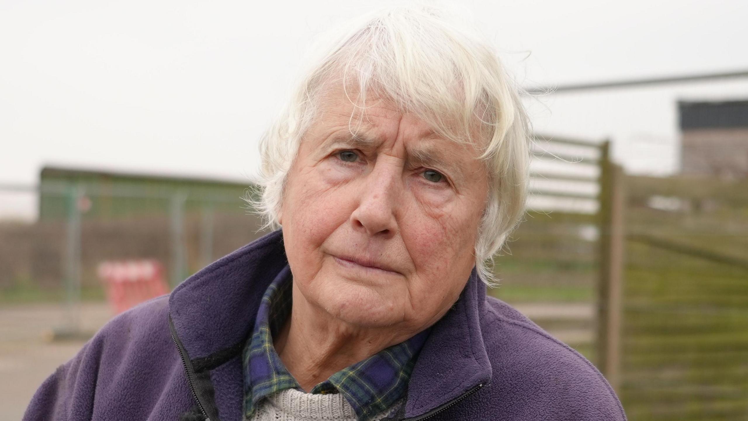 Bryony Nierop-Reading, a woman with short grey  She is wearing a blue and green checked shirt with a cream jumper. She is also wearing a purple fleece and is looking into the camera. 