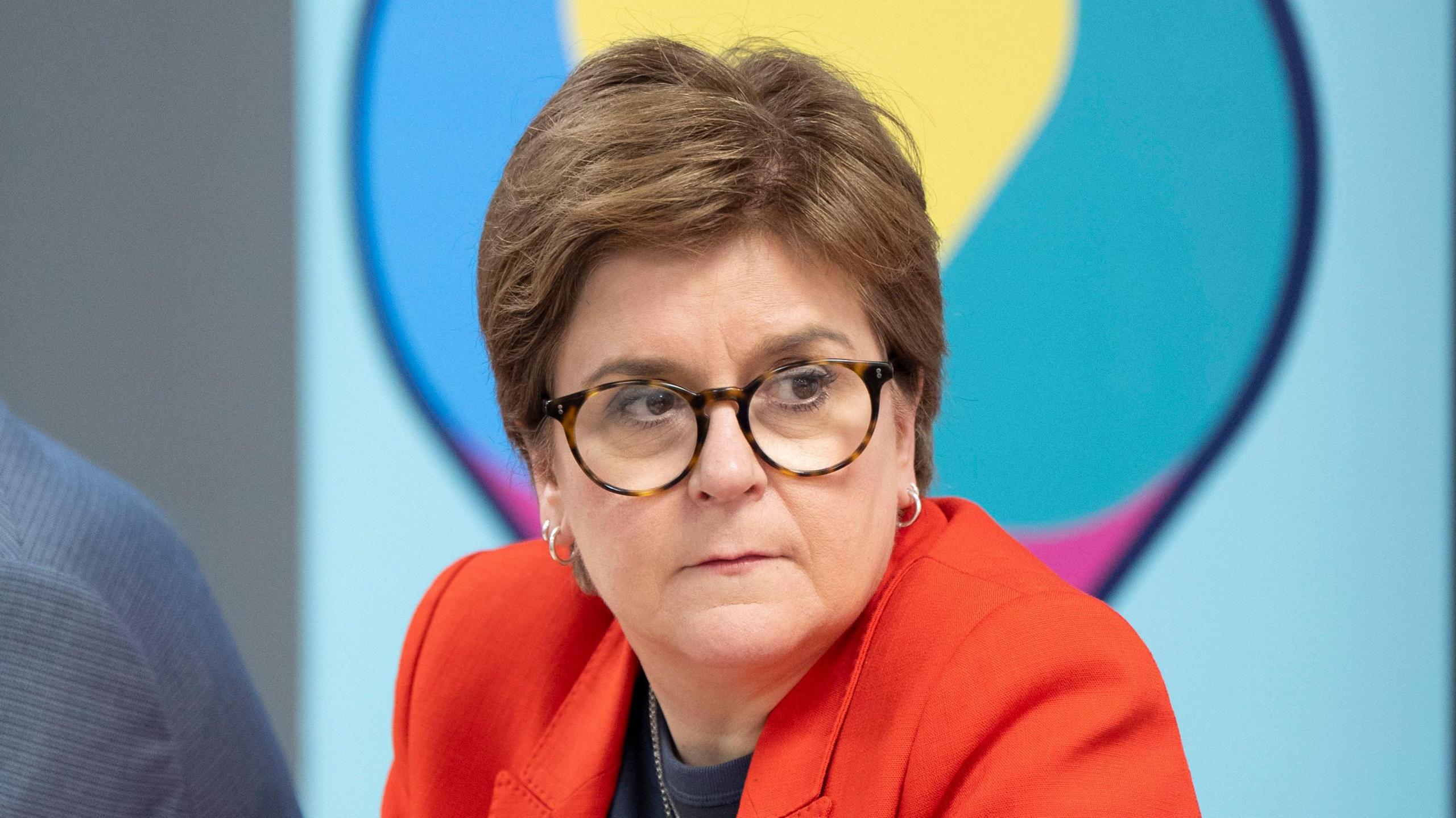 Woman wearing glasses and a red blazer, seated at a table and looking to her left, with a blurred colourful background behind her.