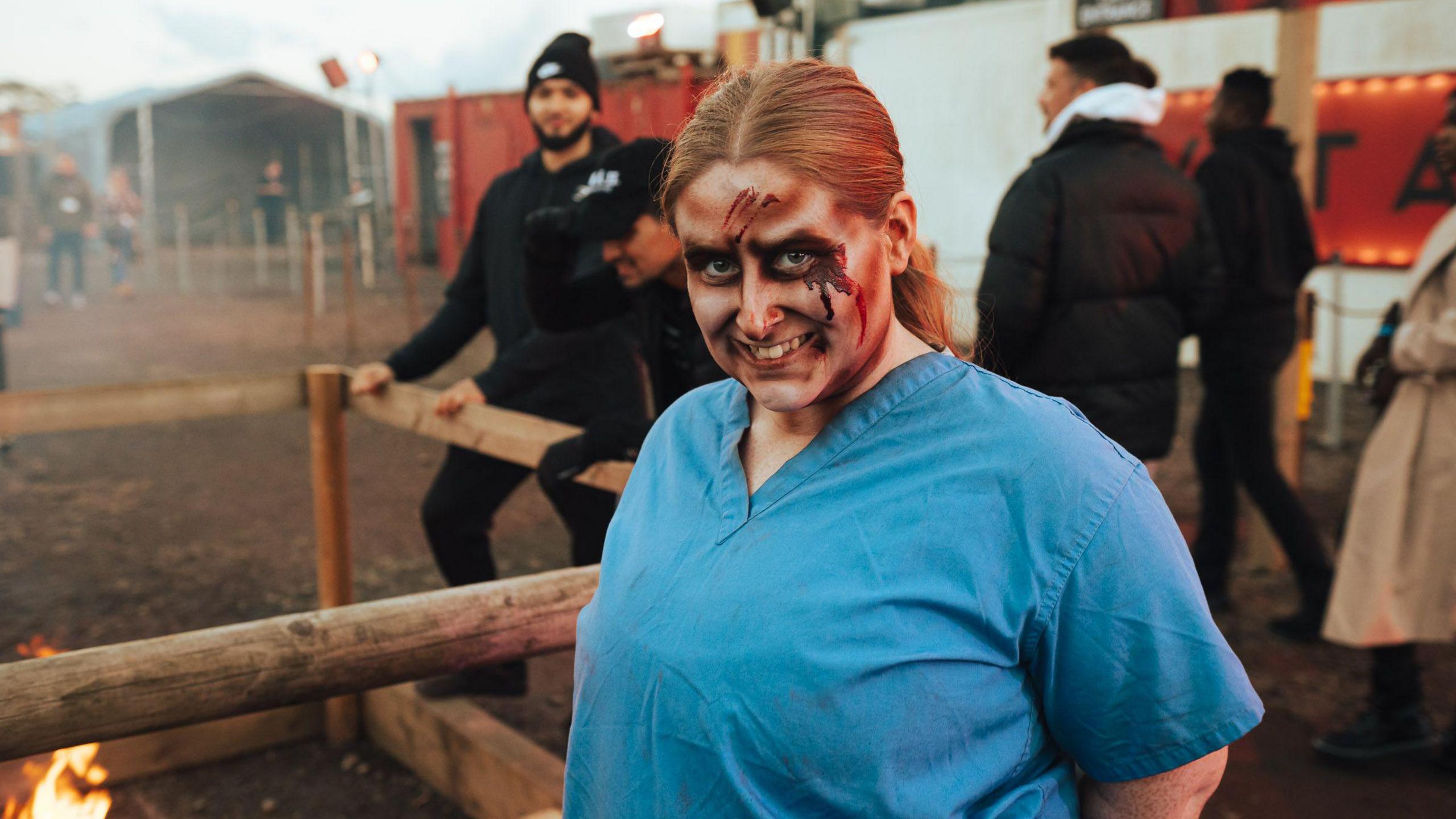 A woman stands next to a wooden pen at Avon Valley Adventure and Wildlife Park. She is wearing blue hospital-style scrubs and has her face made up with Halloween-style make up.