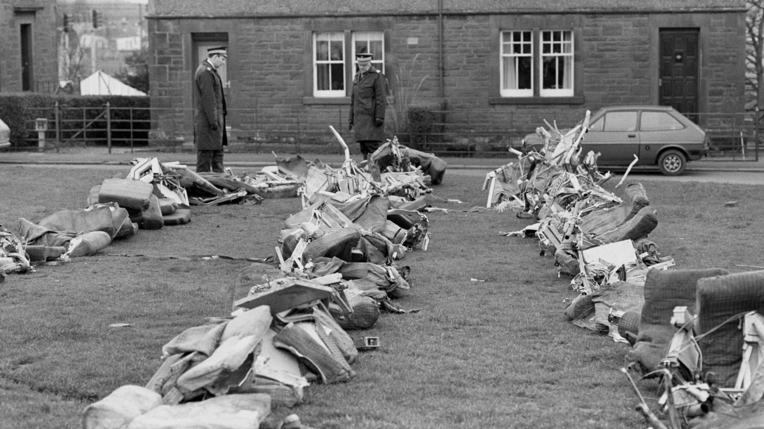 Police survey wreckage from the Lockerbie plane crash. Seats from the aircraft are strewn across grass, with two police officers standing looking at it all. Houses can be seen on the street across from them.