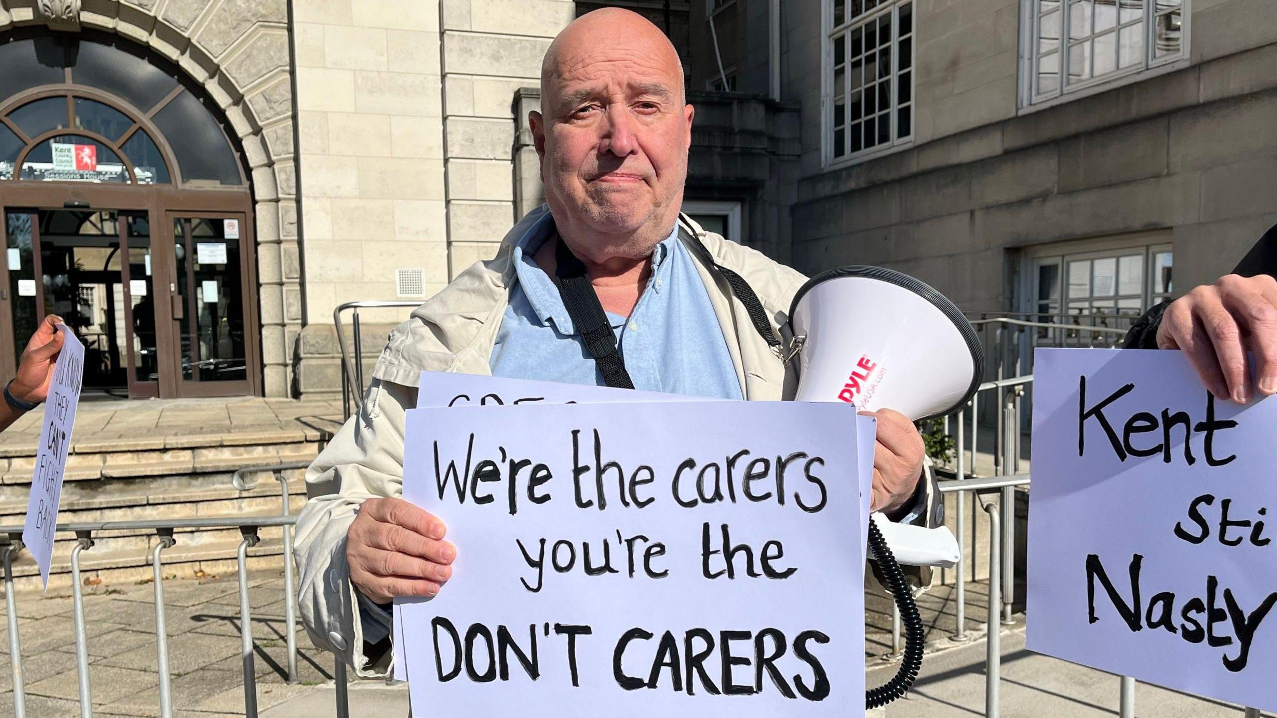 A man in a blue top stands outside County Hall holding a megaphone and a sign which readers "We're the carers, you're the DON'T CARERS"