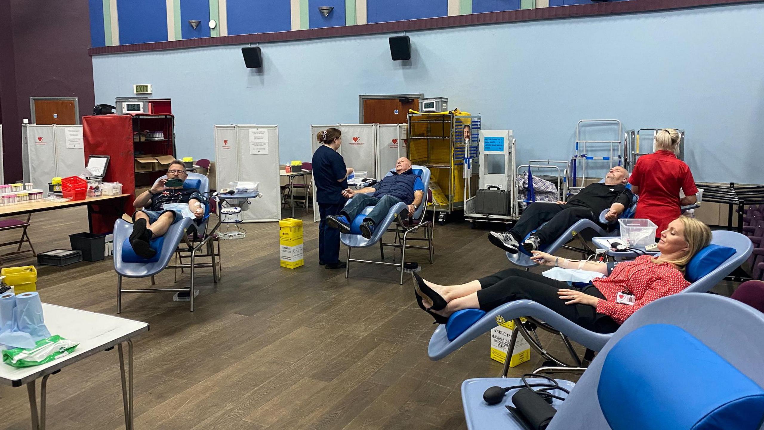 Four school staff members sit back in reclining chairs, with plasters and wires on their arms as they donate blood. Two nurses can be seen in the background preparing for the donations, along with empty vials on a nearby table.