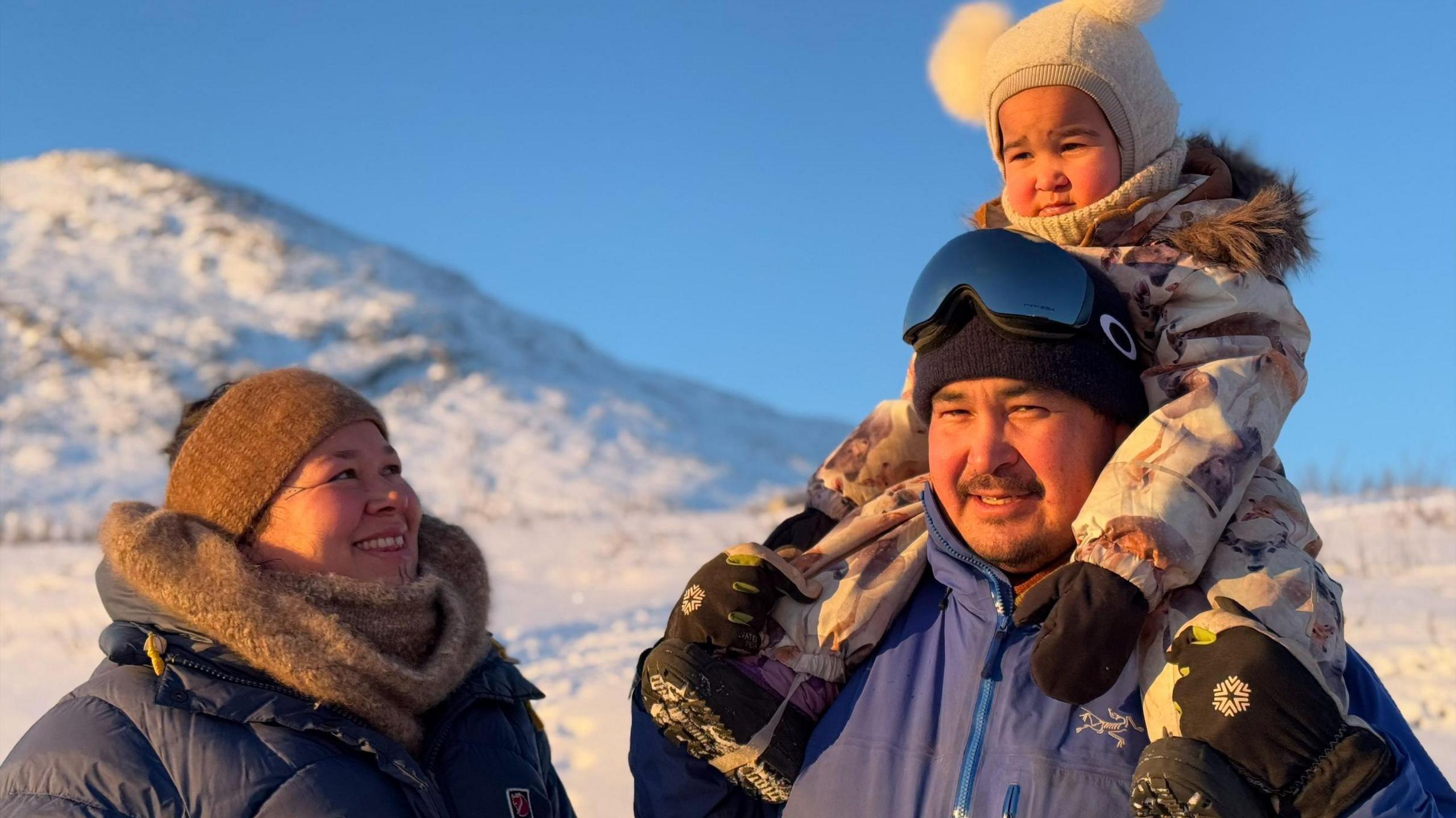 Angutimmarik Hansen wearing goggles and a beanie holding a small child on his shoulders with his wife standing beside him smiling