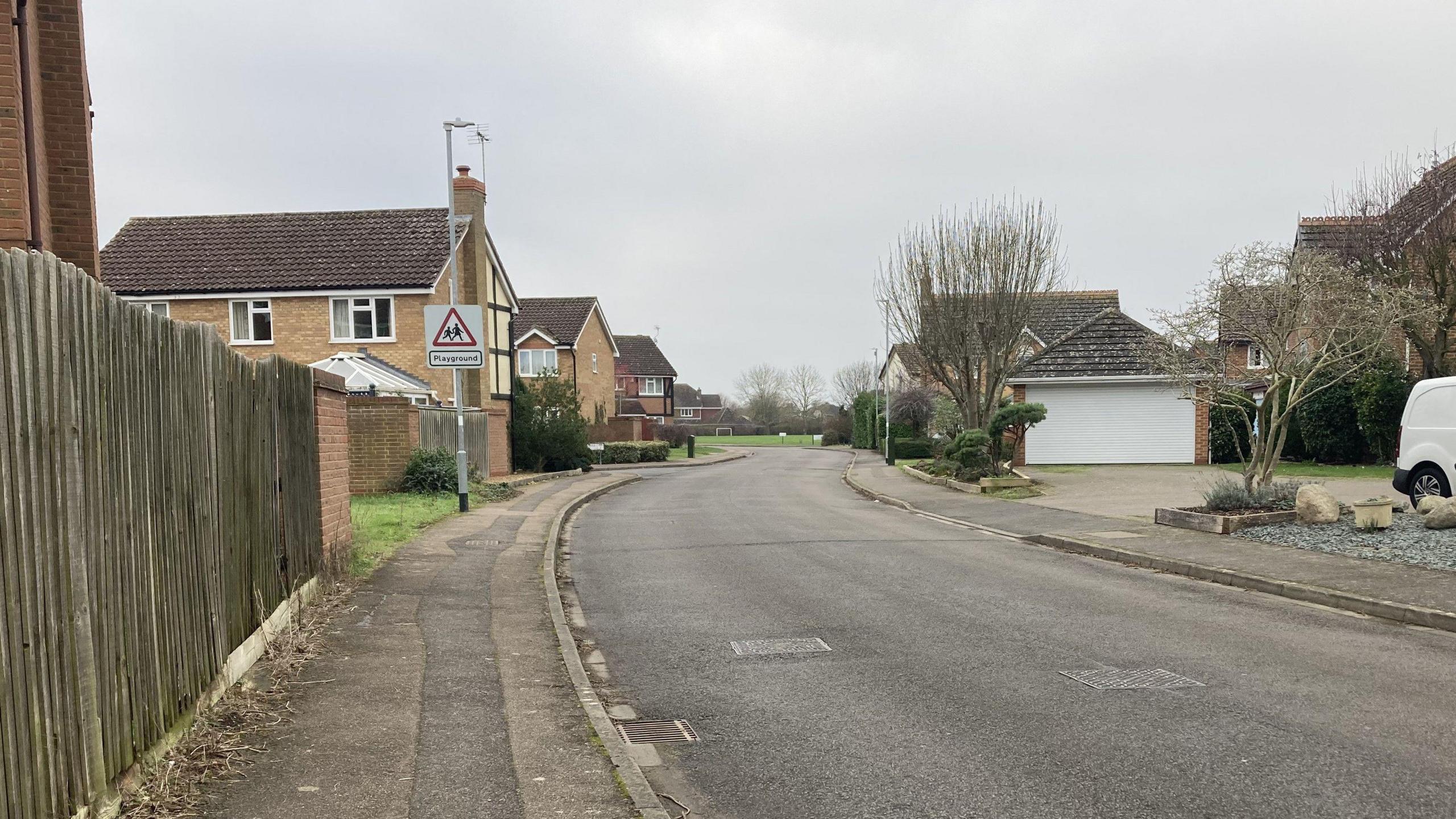 The same street in day time. Houses and bare trees are visible on both sides of the road. A triangle playground sign is on a nearby street light. 