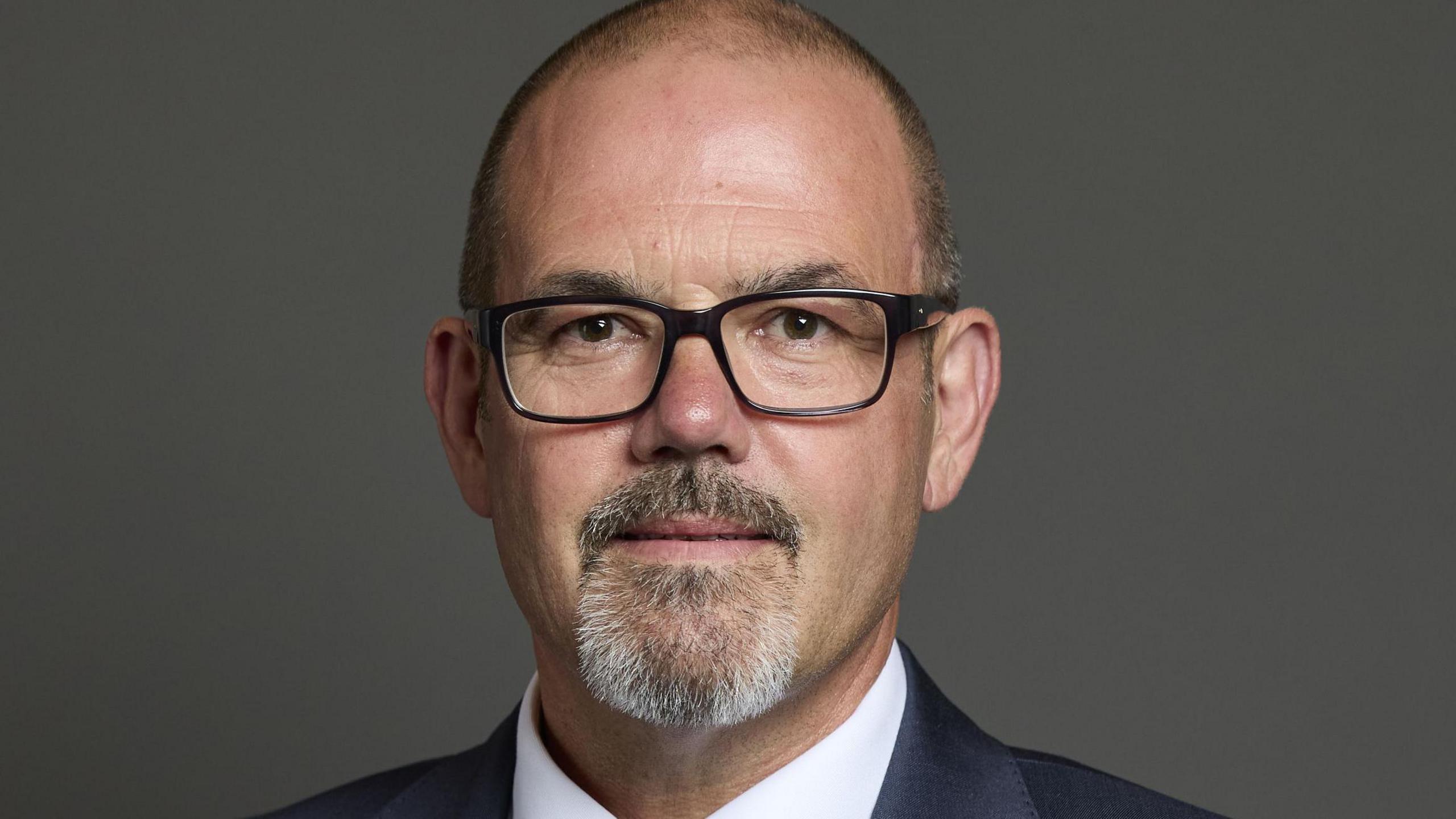 Official portrait of Paul Foster. It's a head and shoulders shot and he is standing in front of a dark grey background. He has close cropped hair and a goatee beard with white in it and black-framed glasses.