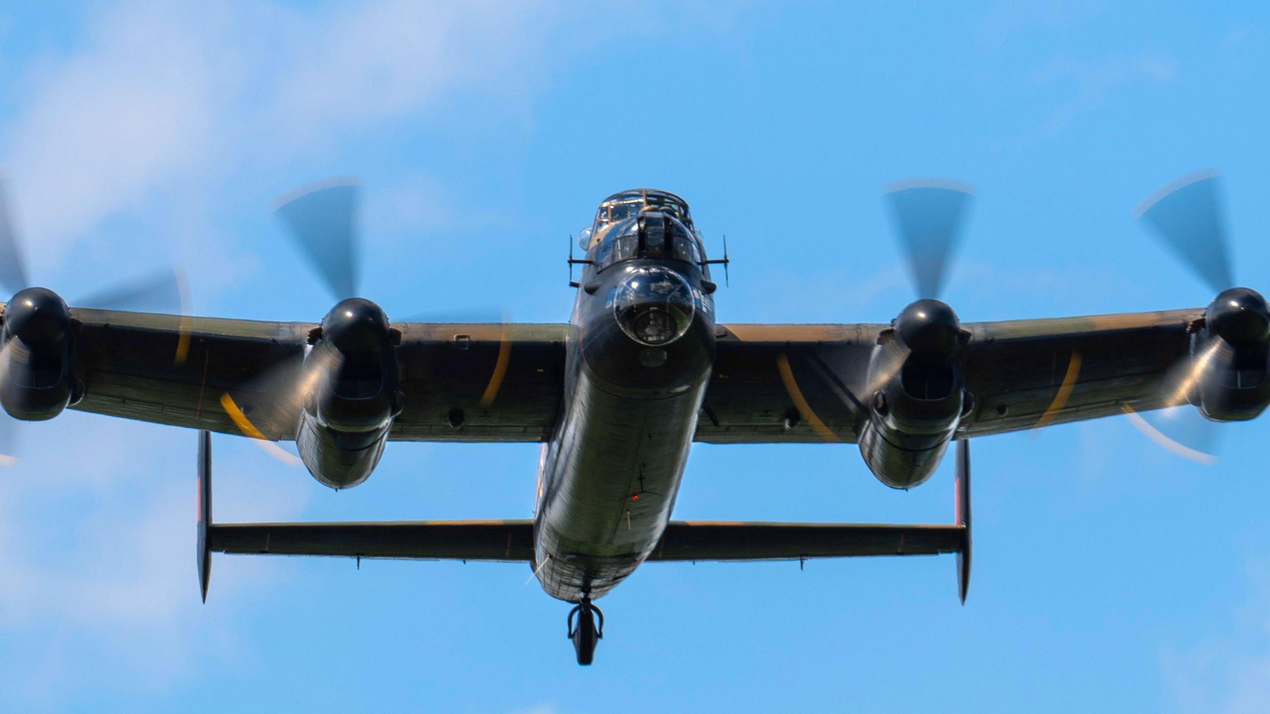 A Lancaster bomber flies overhead. You can see its undercarriage, four whirling propellers and double tailplane