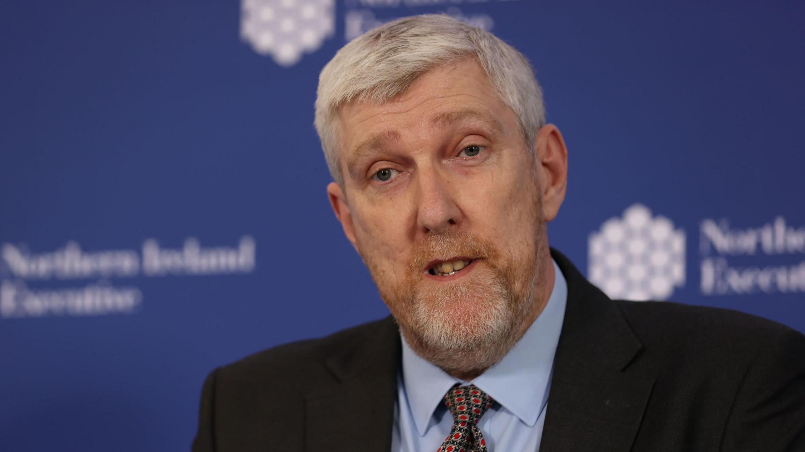 Finance Minister John O'Dowd mid-speech at a Northern Ireland Executive press  event.  He has short, greying hair and a beard.  He is wearing a black suit jacket, a light-blue shirt and a red and blue patterned tie. 