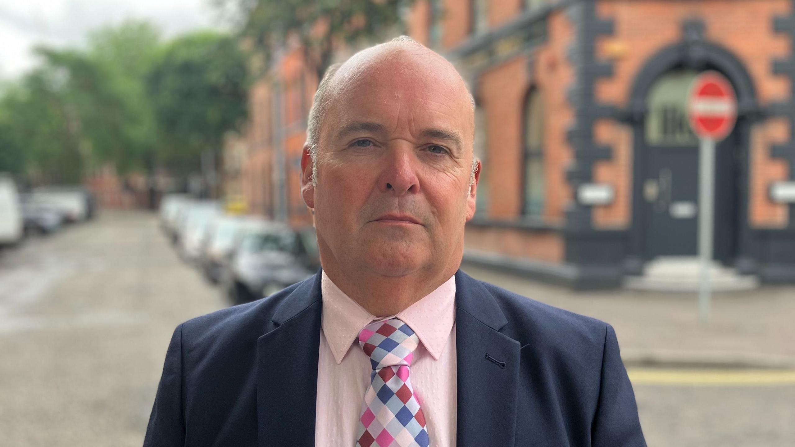 A white man looks directly into the camera unsmiling. He is balding with short grey hair on the sides of his head. He is wearing a navy formal blazer, a pink shirt and a multi-coloured checked tie. The background is blurred but he is standing on an empty street with a red brick building behind him and a number of parked cars. 