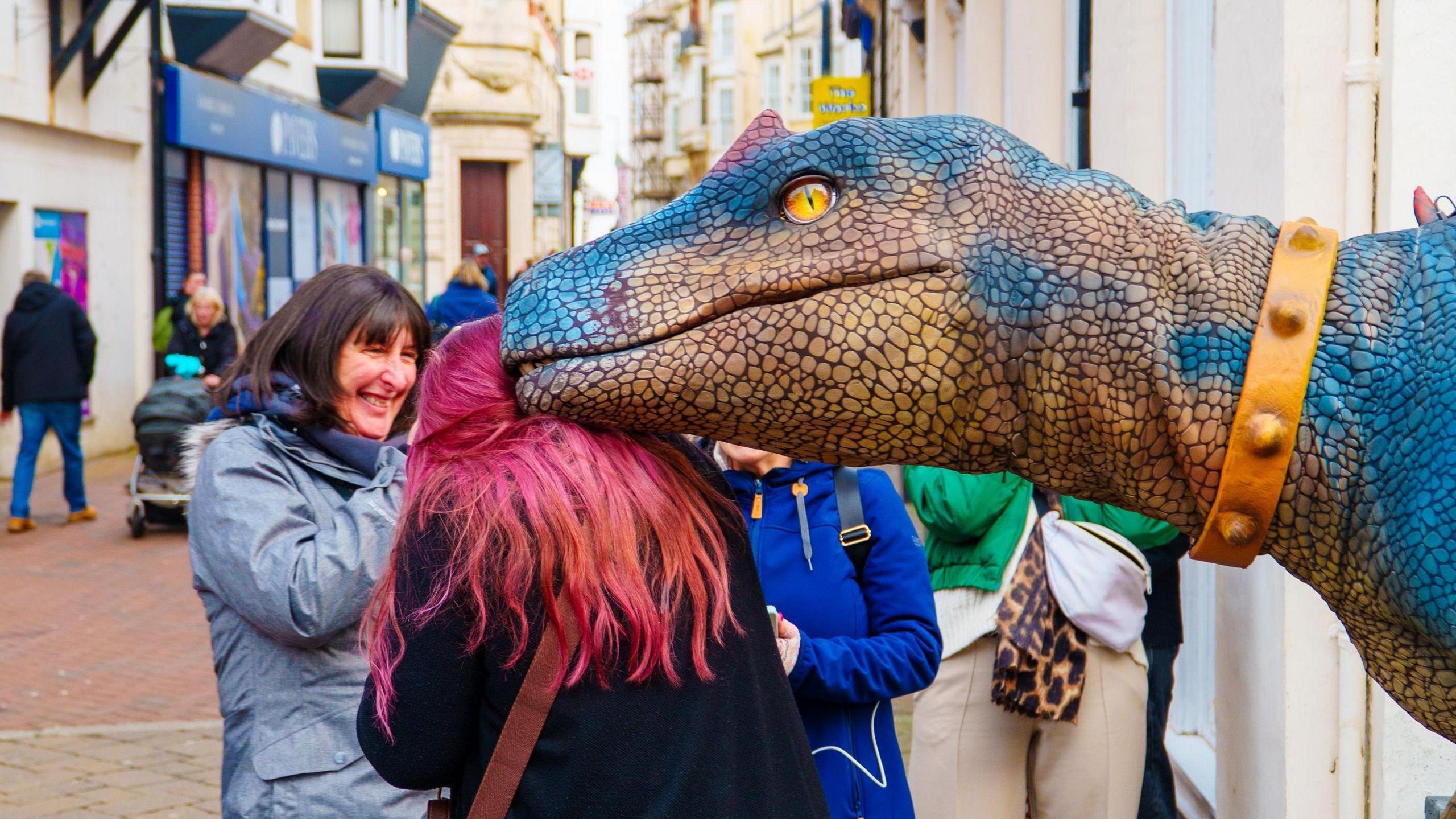 An animatronic dinosaur rubbing its head against the head of a woman with pink hair. A woman is watching and laughing.