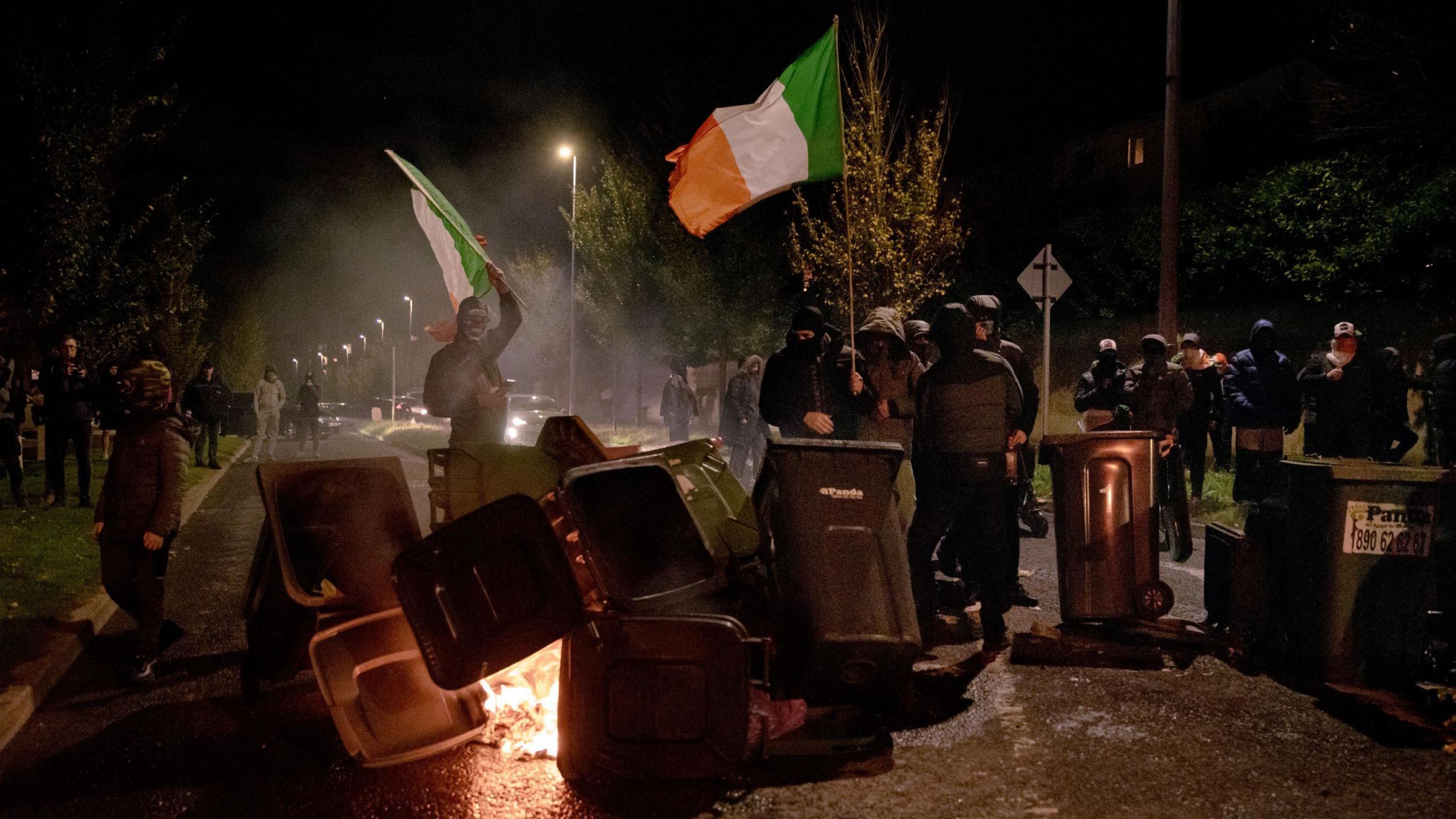 People are dressed completely in black. They are flying two Irish flags. In front of them is a collection of black bins. In between the bins a fire has been started. The sky is dark with street lights in the background.  