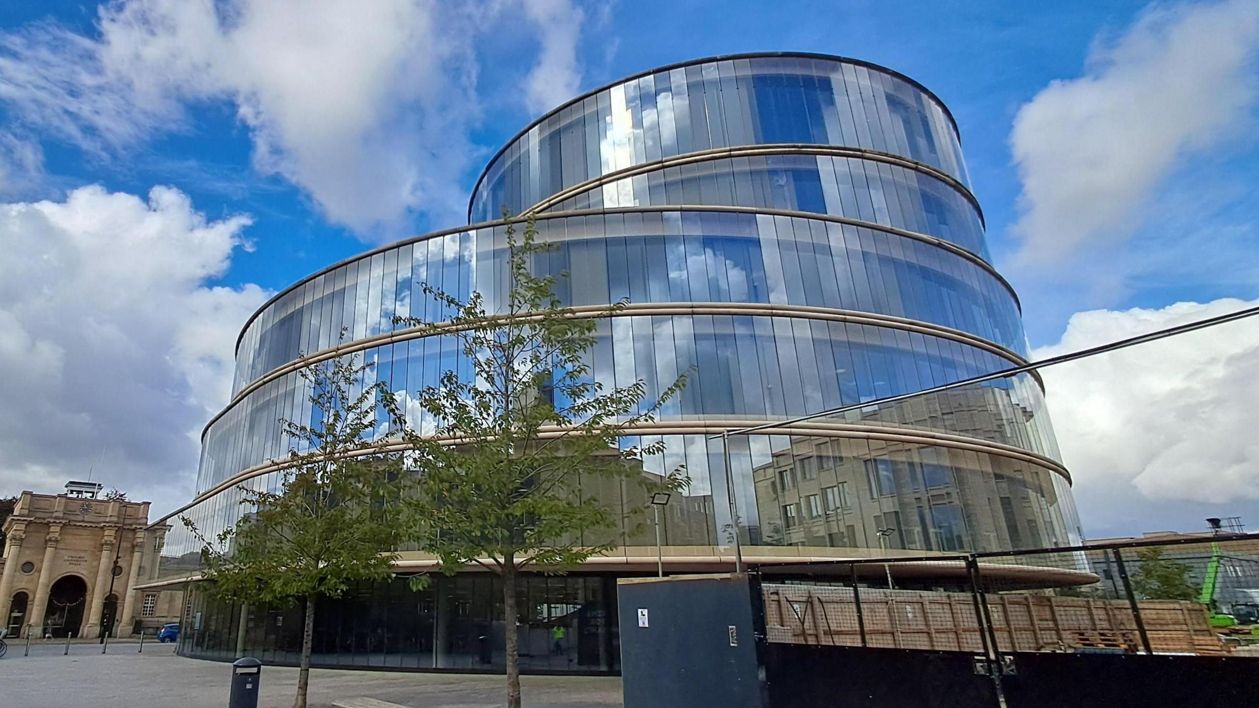 A circular multi-tiered building has reflective glass surface which shows the blue skies and white clouds that hang above it. The building sits in a city street with paving in front of it and a church-like building in the background.