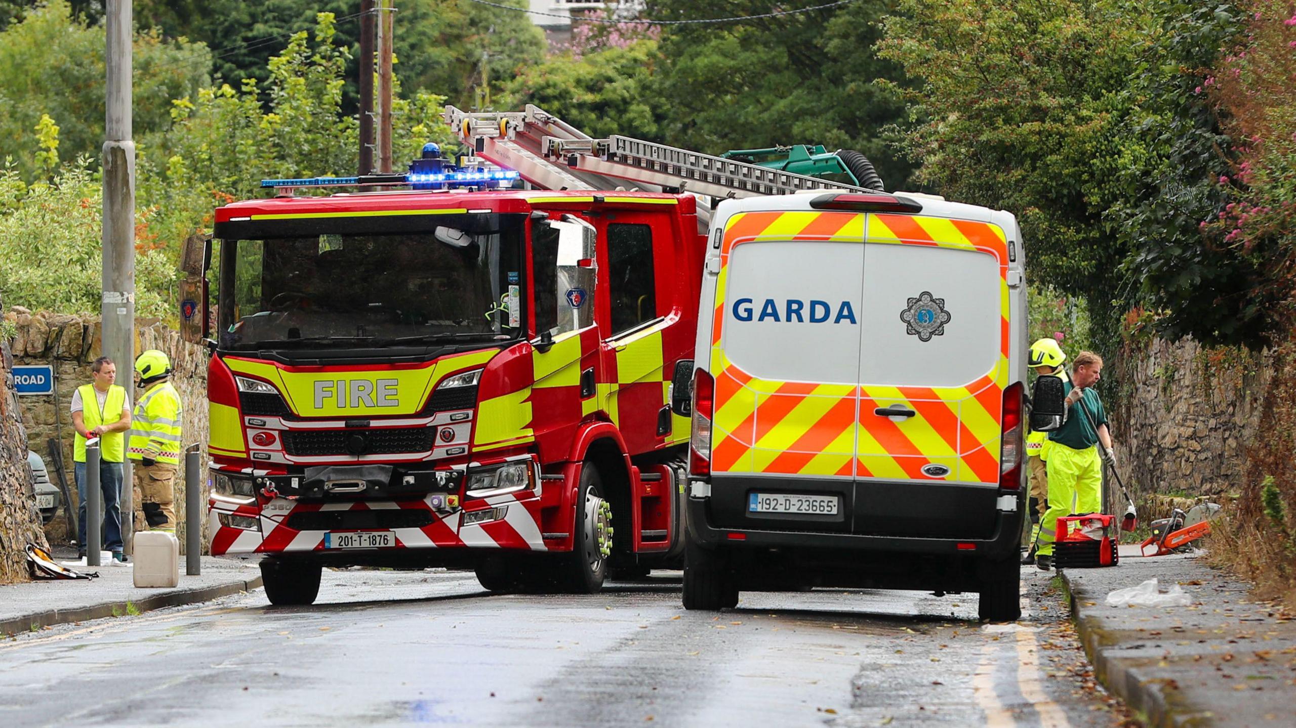 A fire engine and a Garda van parked on a road, with emergency service workers standing on either side. 