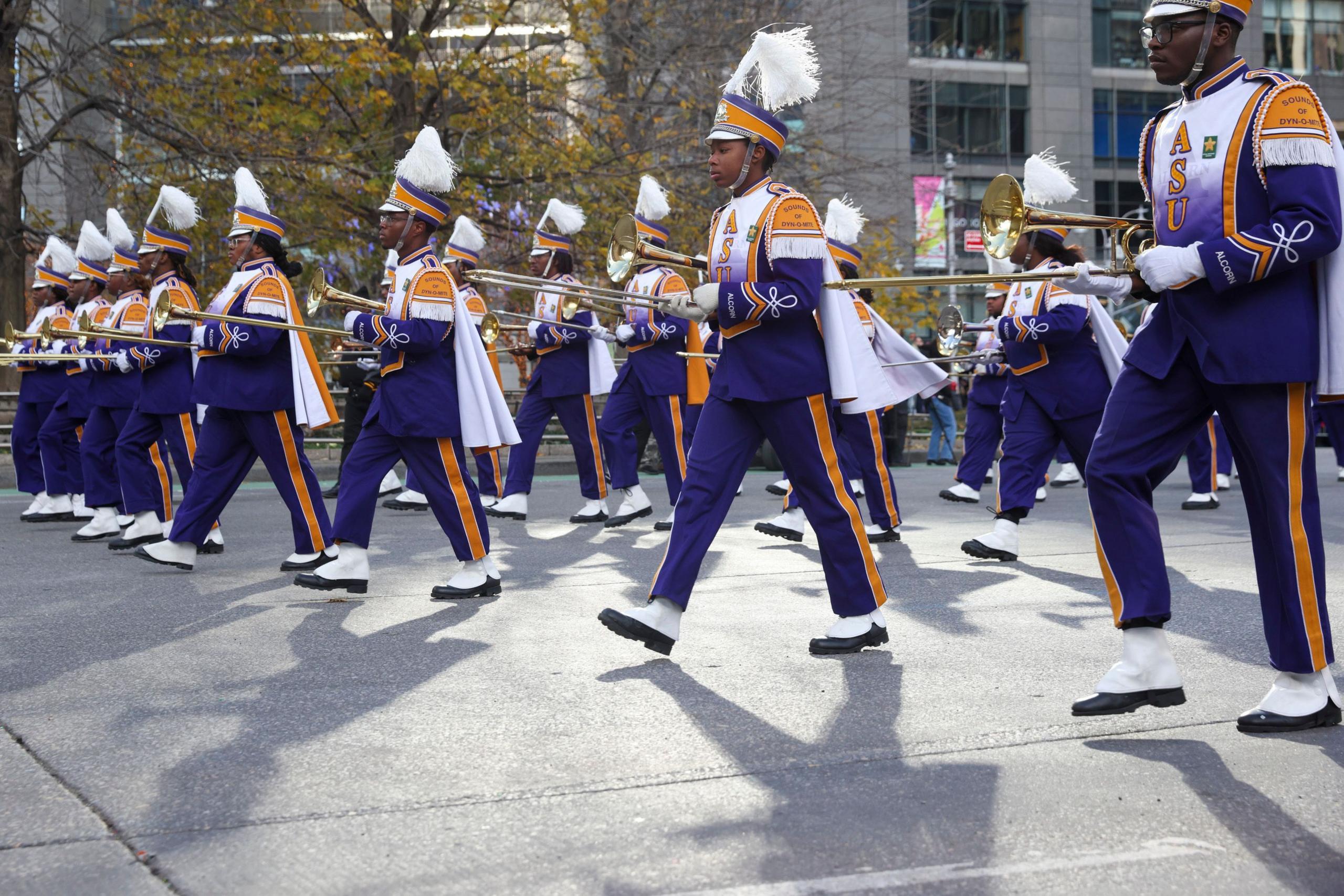 A marching band wearing dark blue, white and gold uniforms marches on the street holding trombones.