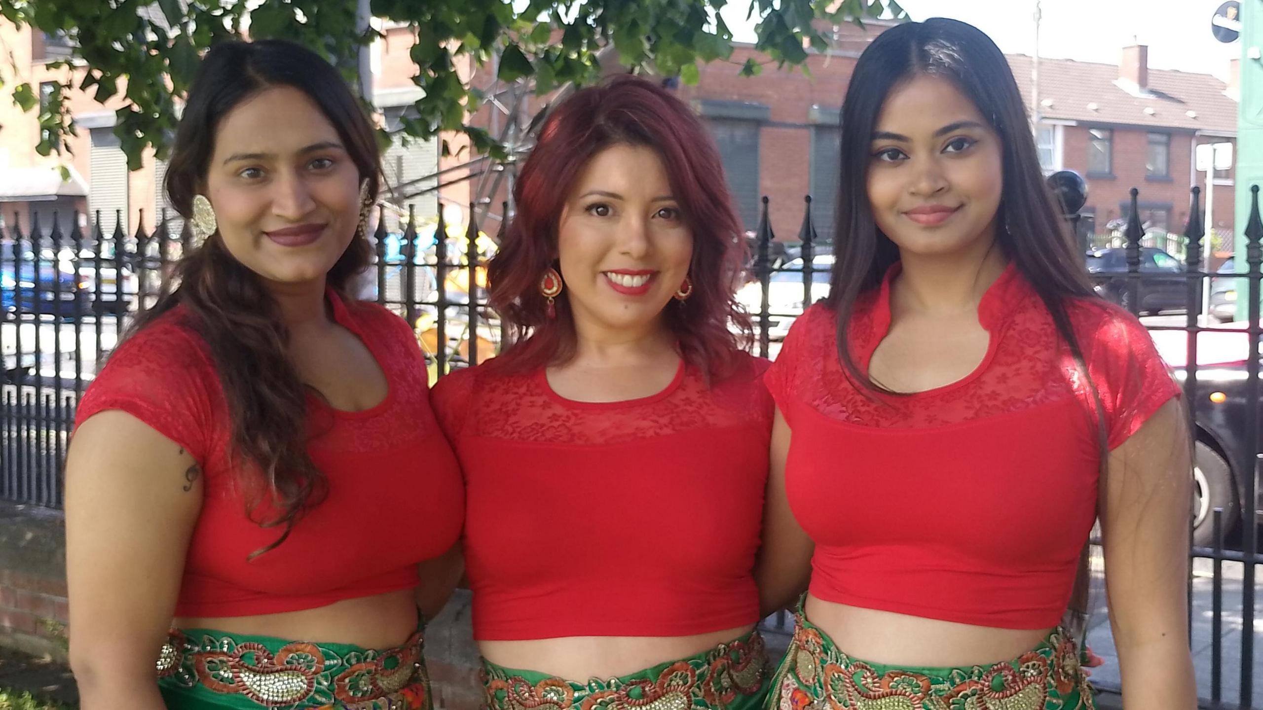 Three women standing, smiling at the camera. They are wearing red tops and green skirts.
