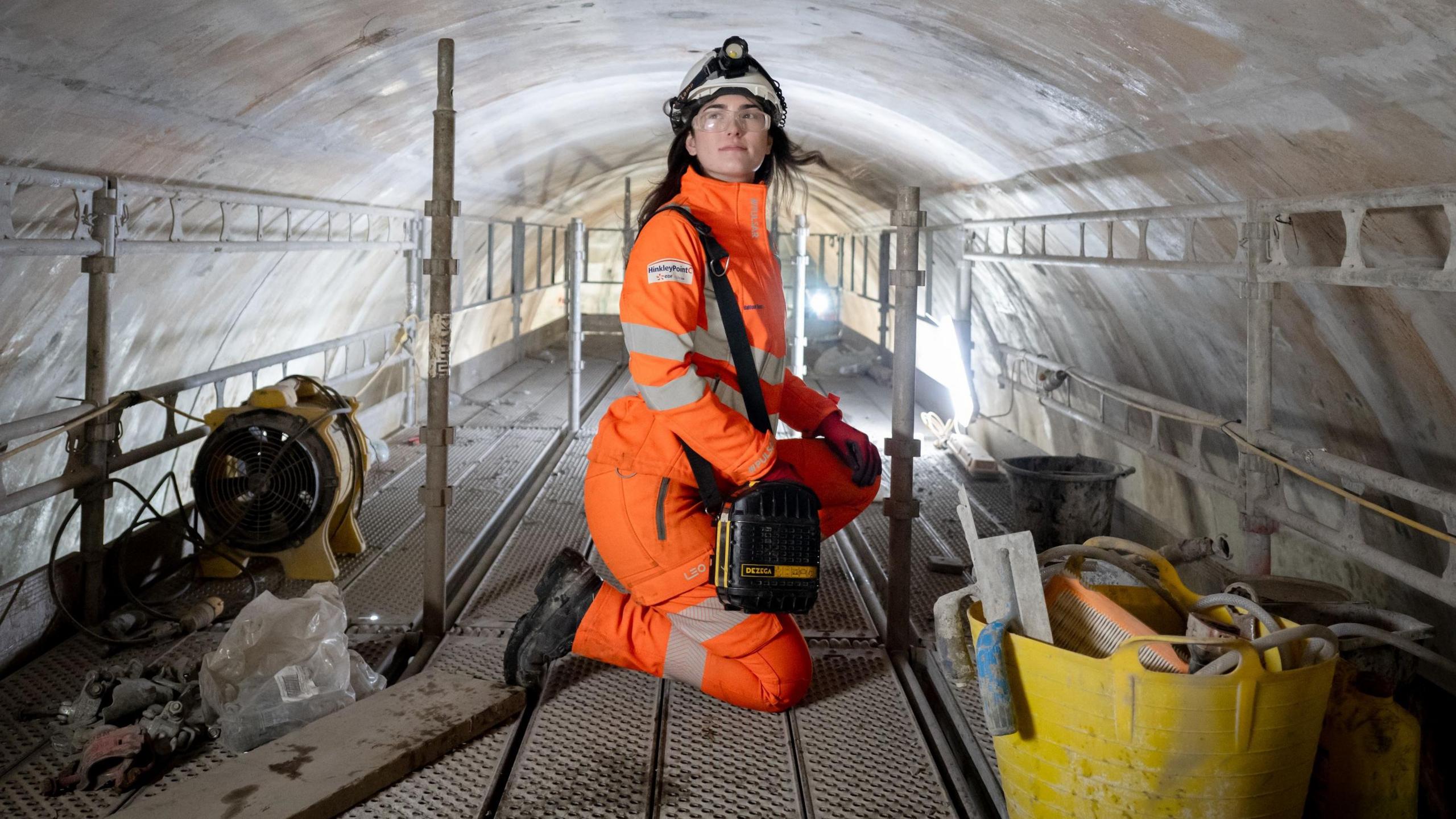 A woman in all-orange overalls, a white safety helmet and clear safety glasses, crouches in a large concrete tunnel