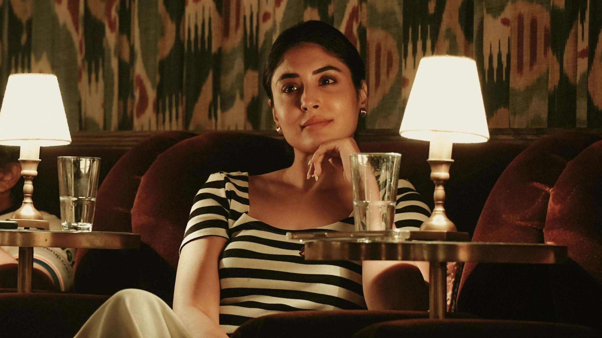 A woman sitting on a maroon sofa wearing a black and white striped t-shirt. A glass of water and a lamp is placed on the table next to her.