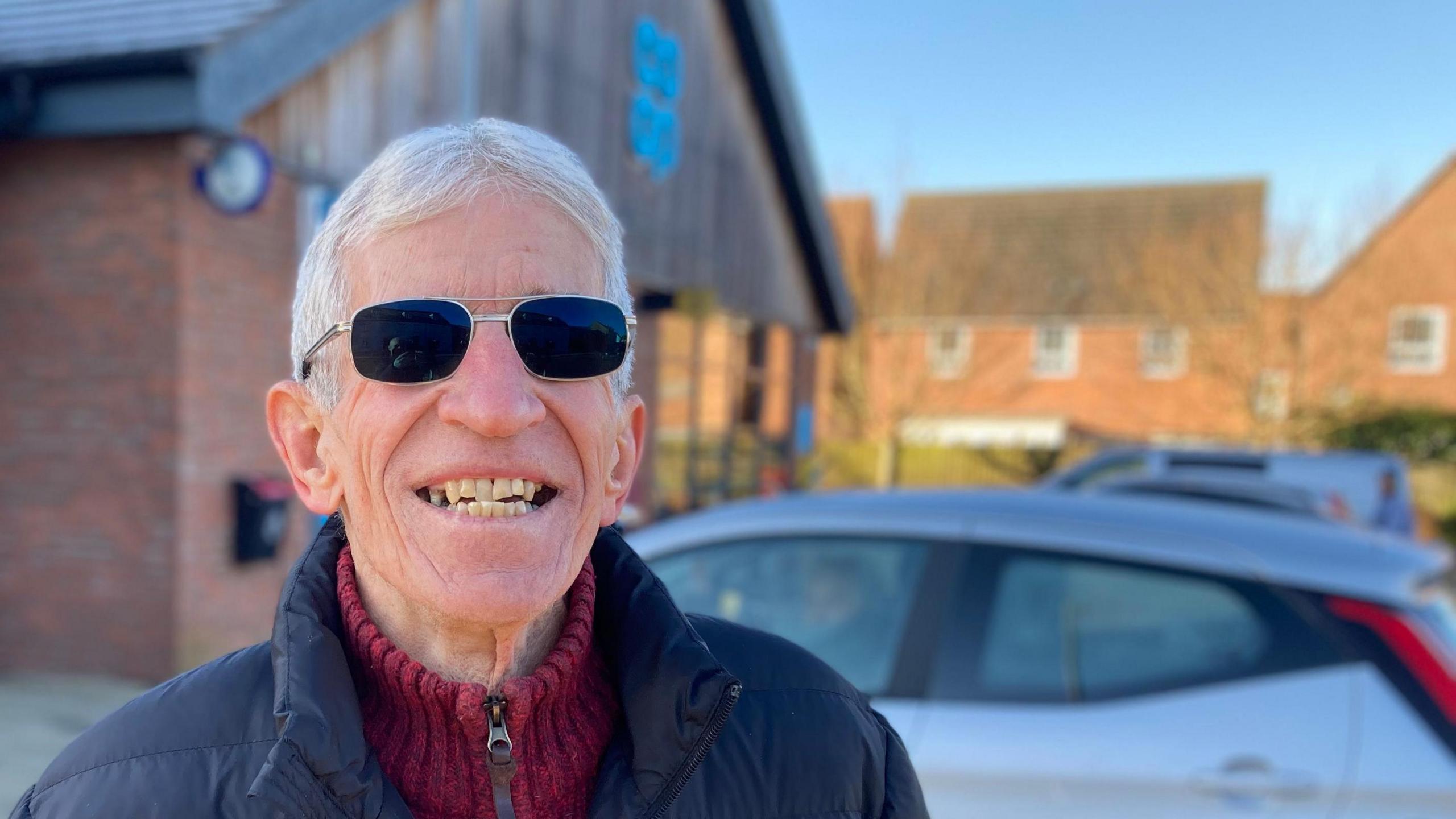 A smiling Mike Main looking towards the camera. He has short grey hair and is wearing a blue padded jacket and sunglasses.  