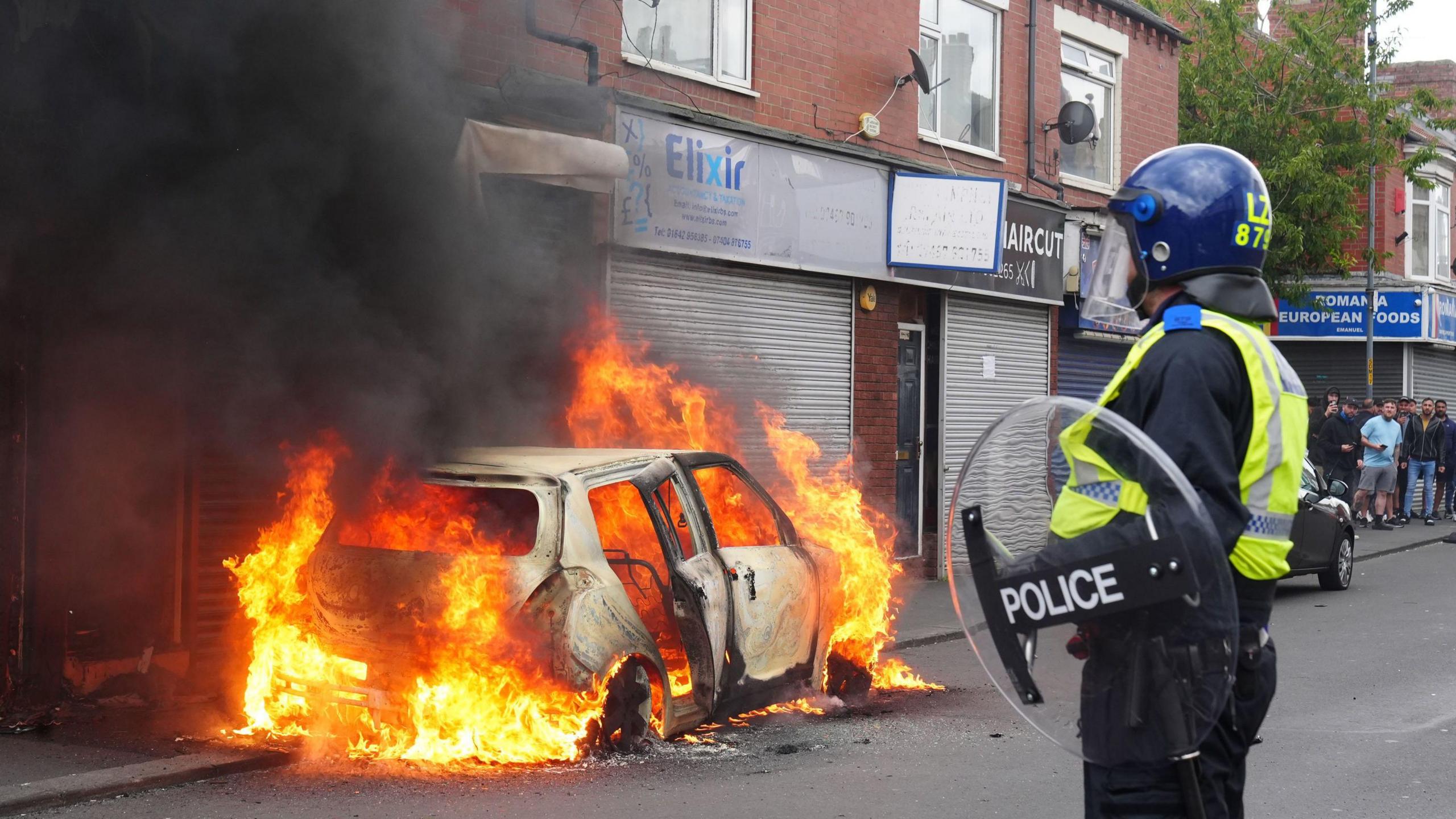 A car burns in Middlesbrough as a police officer looks on