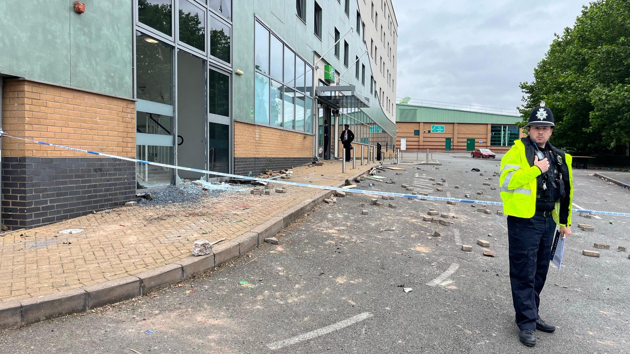 A police officer stands in front of blue and white police tape with his hand on his radio. Behind the tape is a hotel building, with its windows smashed, and rubble strewn across the road in front of it.