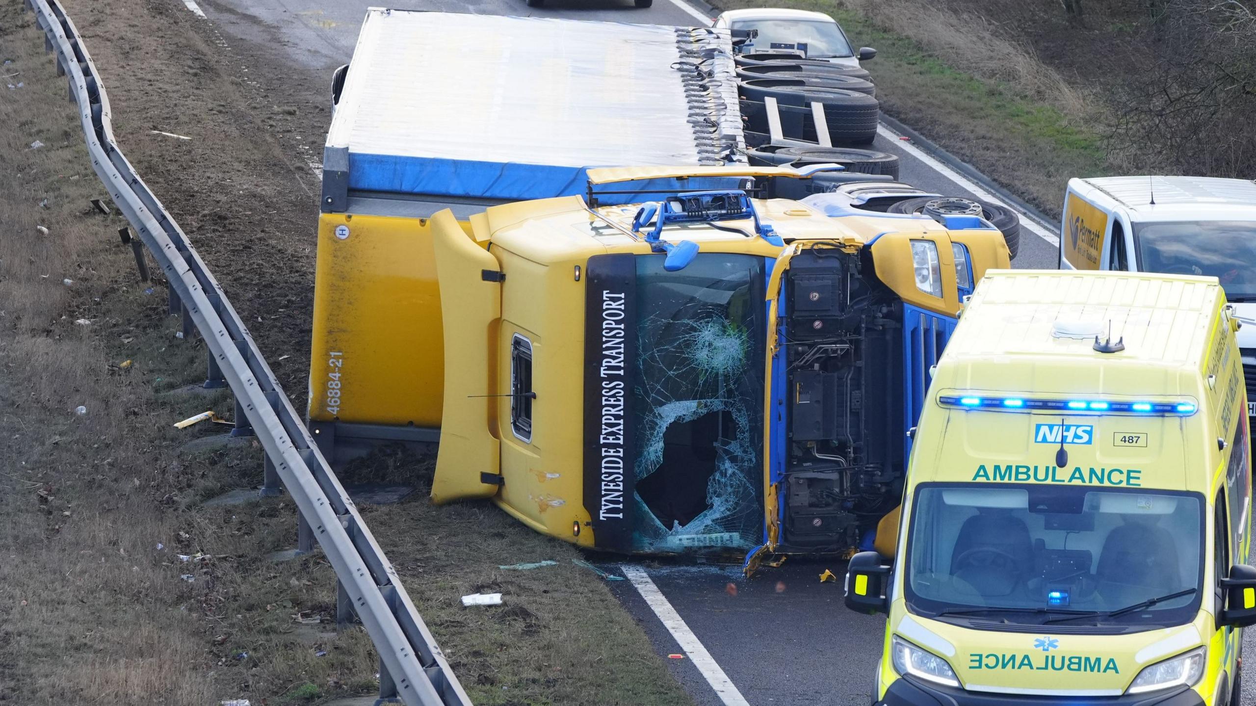 A lorry on its side with a smashed windscreen. There is an ambulance parked in front of it.