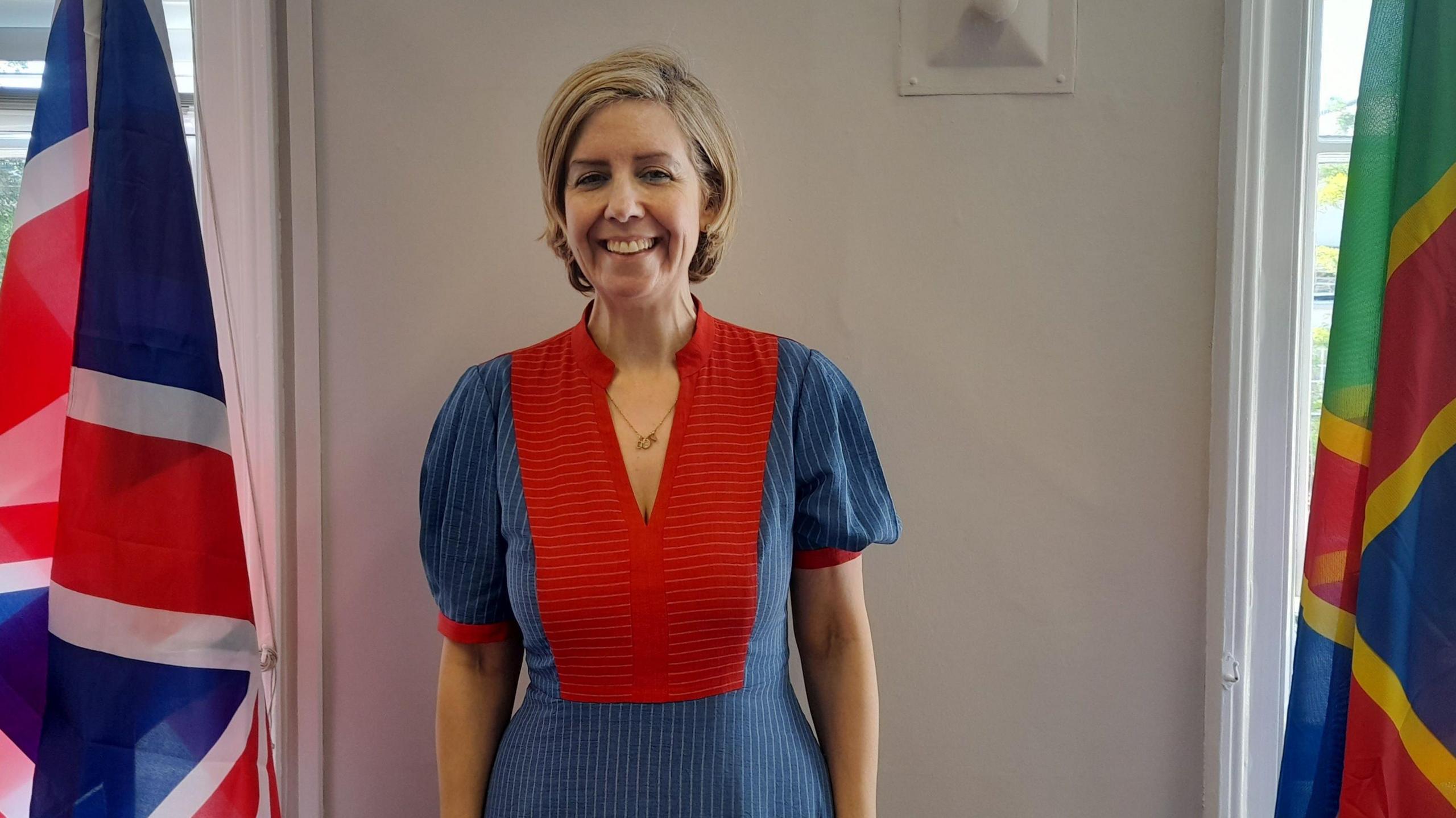 A woman stands between two flags. On the left is the Union Jack, the national flag of the United Kingdom. On the right is a multi-coloured flag featuring green, yellow, red, and blue. She is wearing a blue and red dress with pin stripe detailing. Behind her is white walls are light-colored walls and windows.