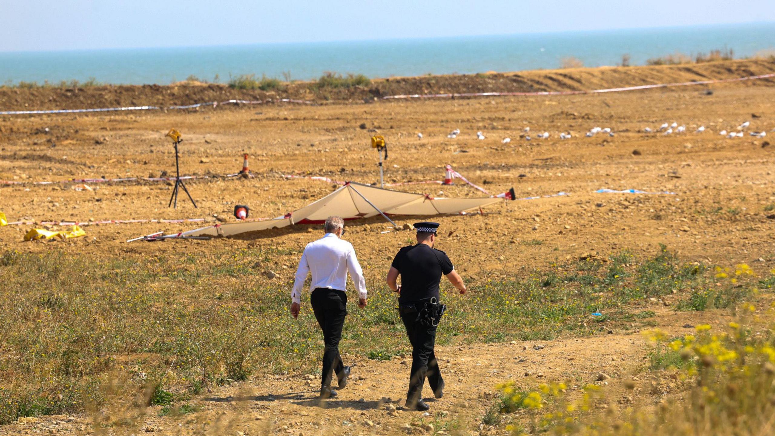 A police officer and a man in a white shirt walk towards the crime scene, which on a dune just off the beach.
