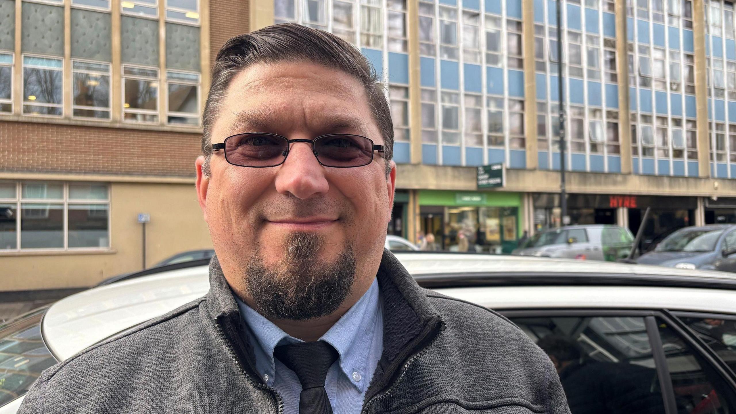 Daniel Pop, pictured standing in front of a car on a busy street in Bristol. There is a large office block behind him. He is wearing a light blue shirt, a dark tie, a grey fleece and dark glasses He has brown hair, swept over and back to one side, and a goatee. 