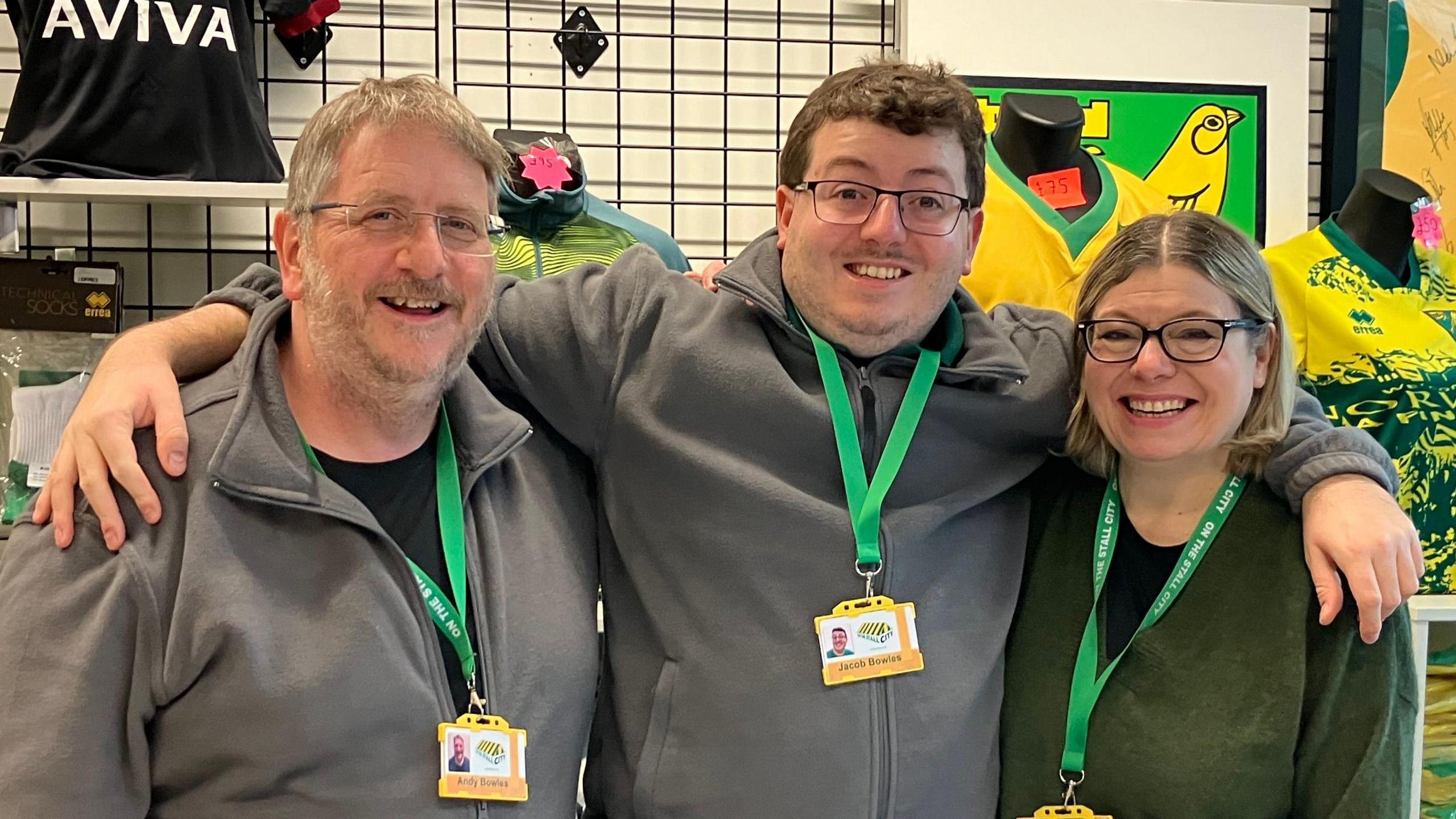 Members from the On The Stall City shop standing in a row looking directly at the camera and smiling. Andy Bowles is on the left dressed in a black T-shirt and grey zip up sweatshirt. He is wearing a pair of glasses and has a green lanyard round his neck. 