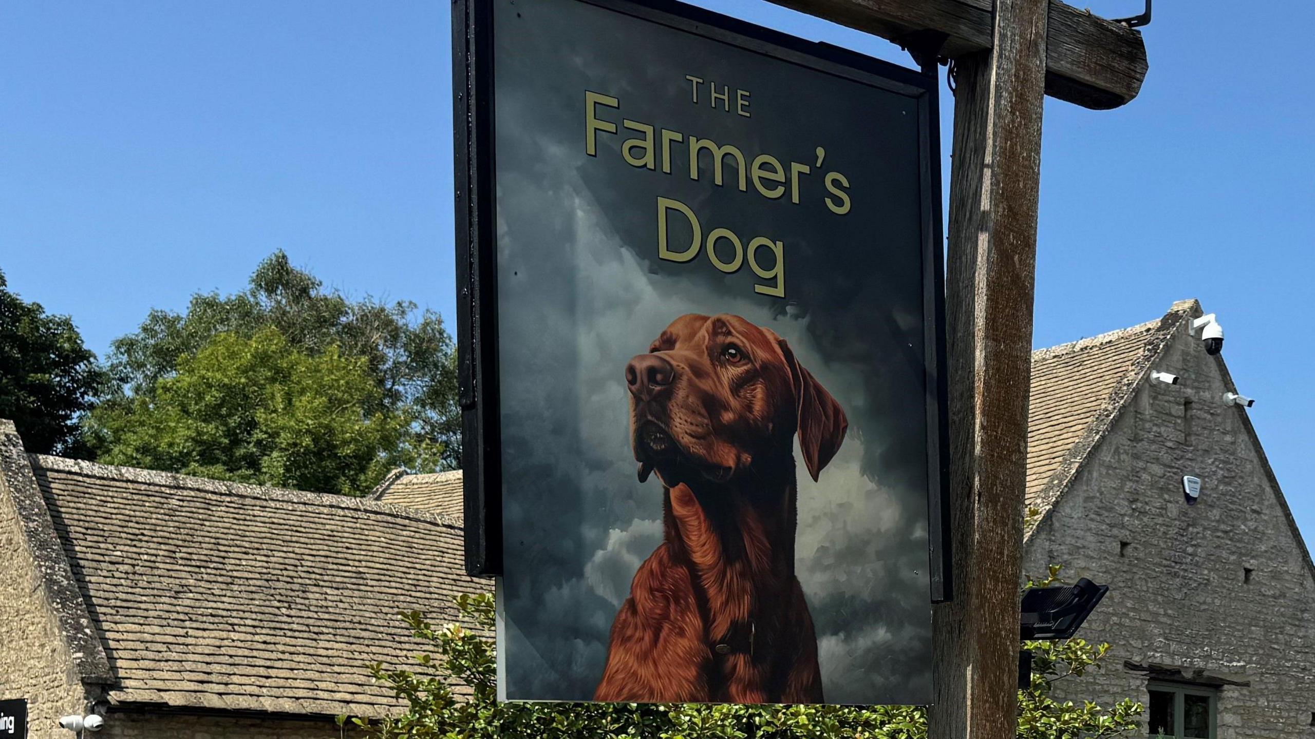 An historic pub under blue skies with rolling fields in the distance and a pub sign with a dog in the foreground
