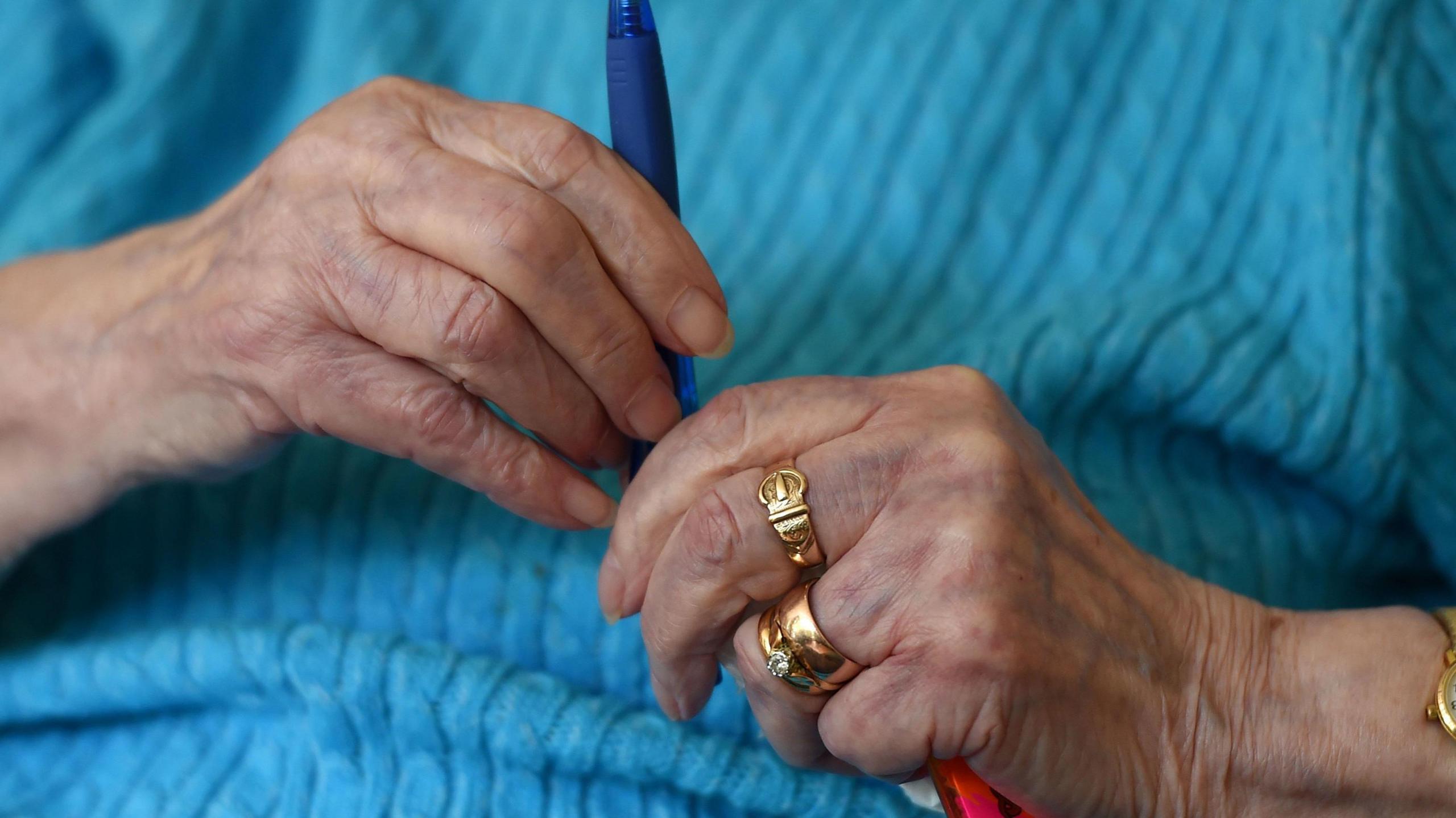 The hands of an elderly woman wearing a blue jumper. She has gold rings and a gold watch on and is holding a pen