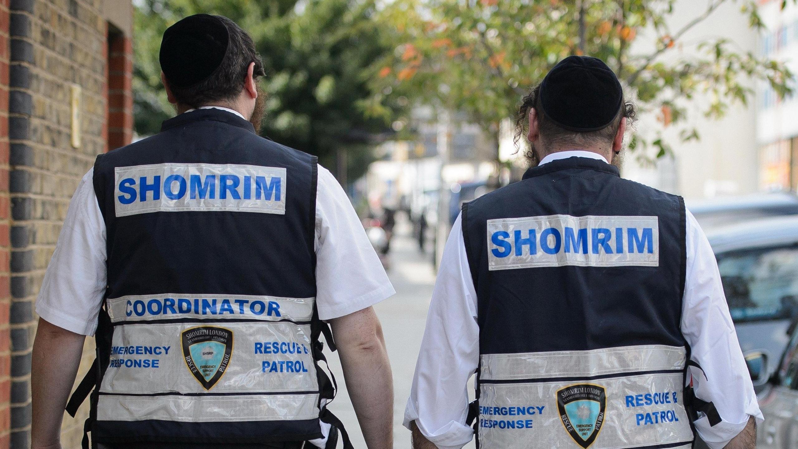 The back of two Jewish men wearing vests labelled Shomrim across the back walk through north London in 2014. They are wearing white shirts underneath the vests and kippahs.