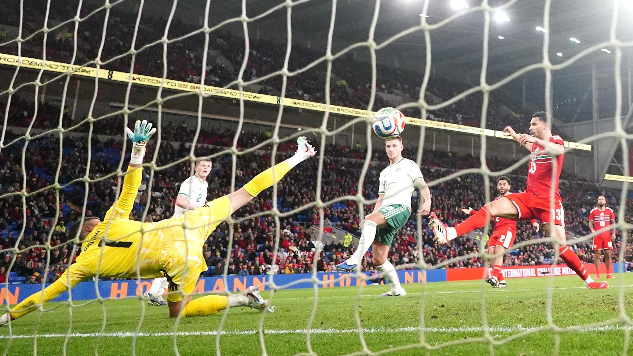 A keeper tries to save a goal scored by Jamie Donley, who is in the centre wearing green shorts and white top. Players from Wales wearing red can be seen in the background.