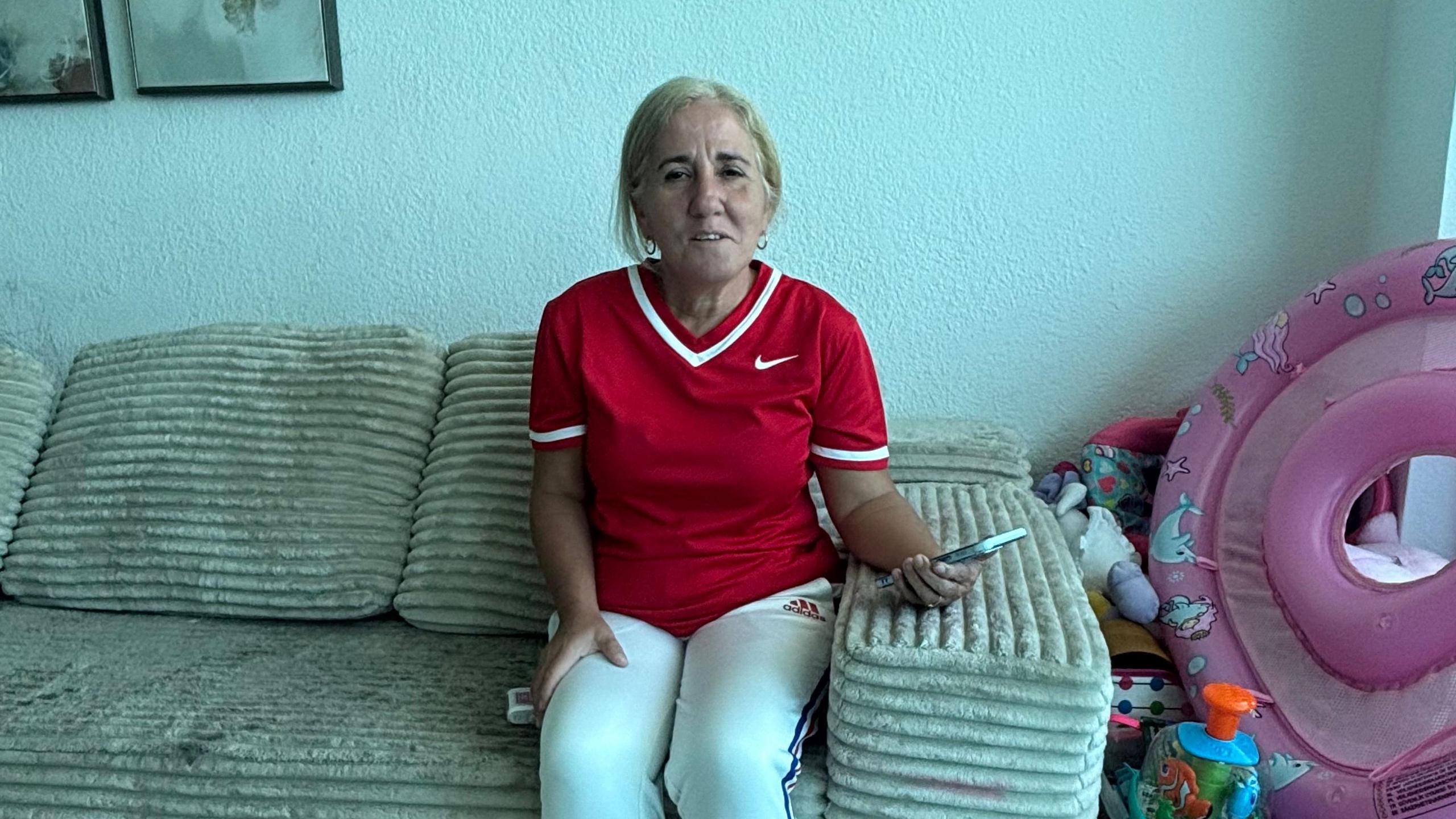 An older woman in a red Nike soccer jersey and white Adidas track pants sits on a beige, worn sofa in an apartment holding her cell phone