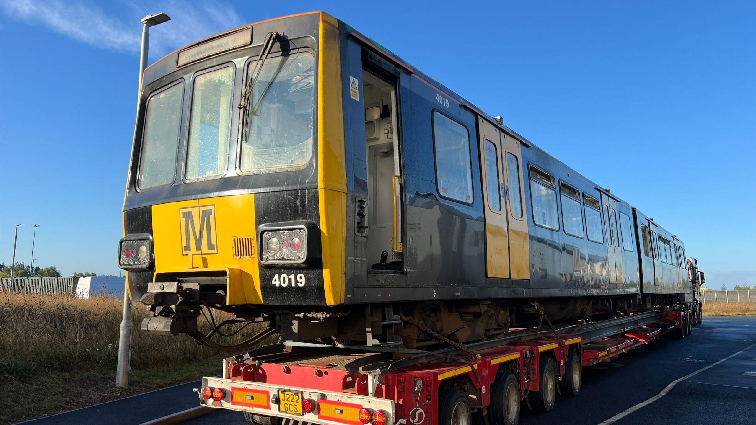 A yellow and black old style Tyne and Wear Metro carriage on a wheeled platform on a tarmac path. It is a sunny, blue sky clear day.