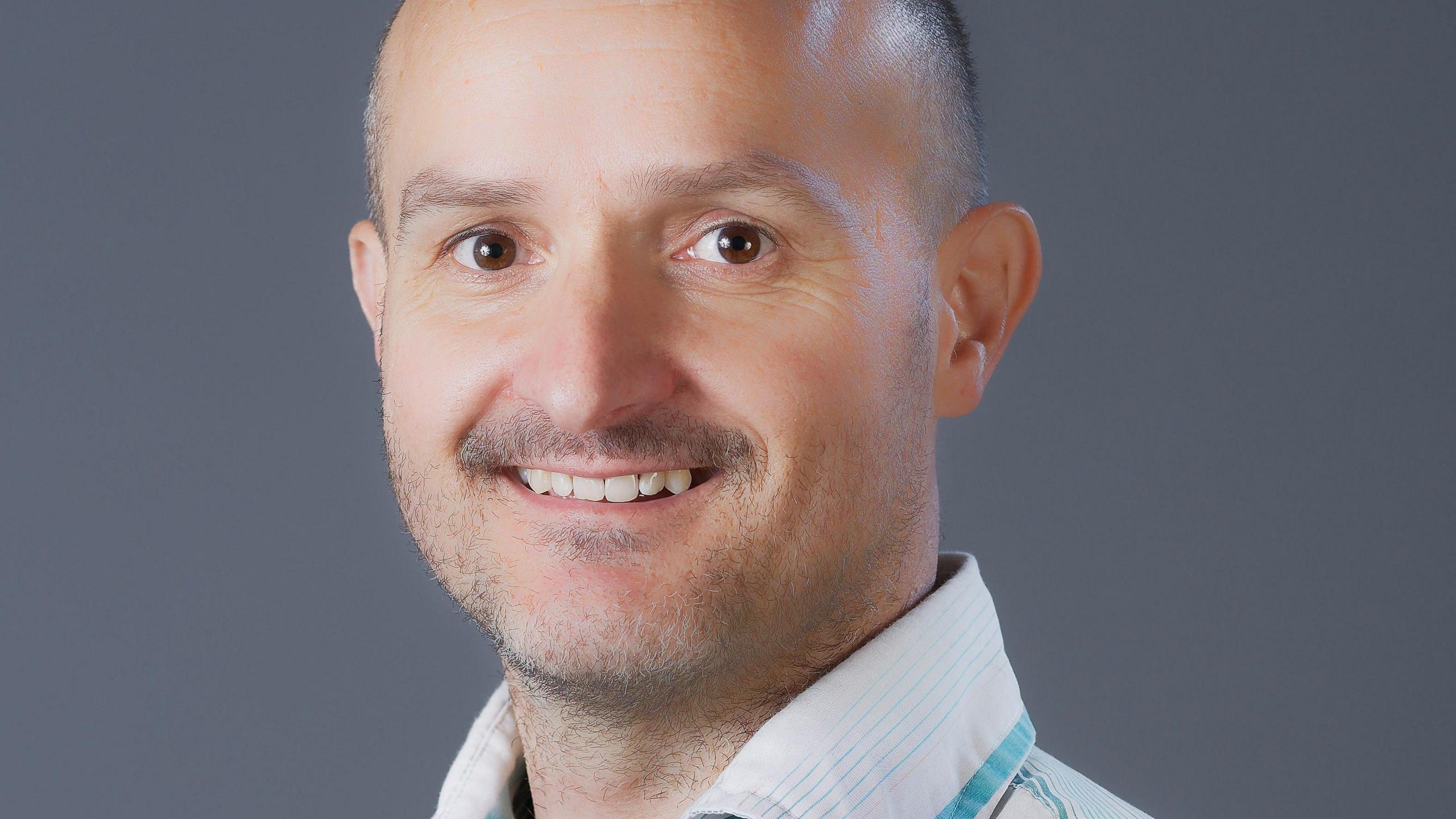 A head shot of Clubb. He is bald on top and has short clopped hair and the sides and a light beard. he is smiling and looking at the camera.